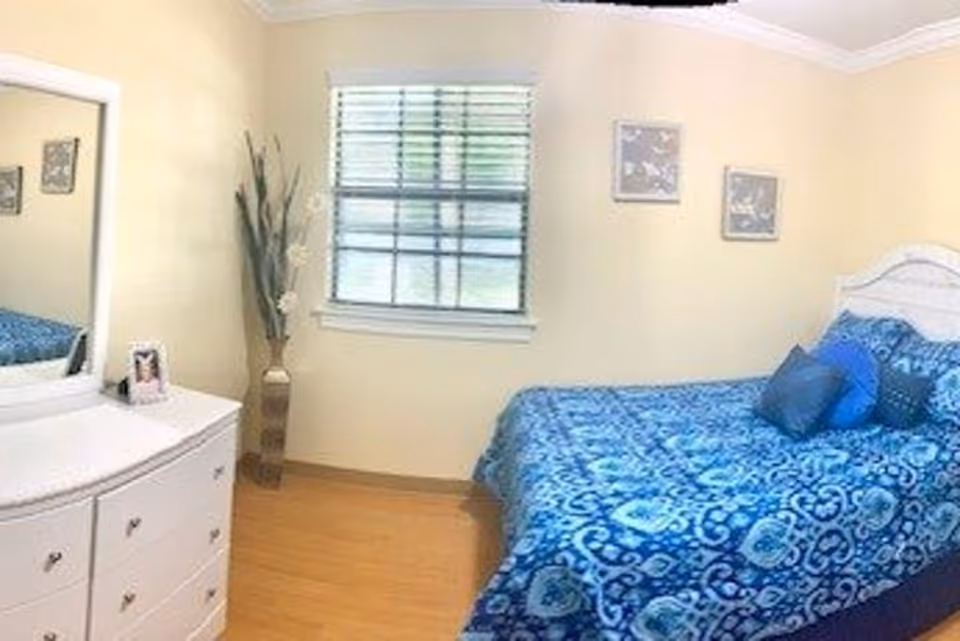 Small bedroom with a blue patterned bed, white dresser and mirror, window, and a tall vase with decorative stems.