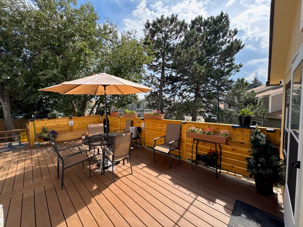 Outdoor wooden deck area with a round metal table and four chairs under a large beige umbrella. The deck is surrounded by a wooden fence with flower pots and planters attached. Trees and neighboring buildings are visible in the background under a partly cloudy sky.