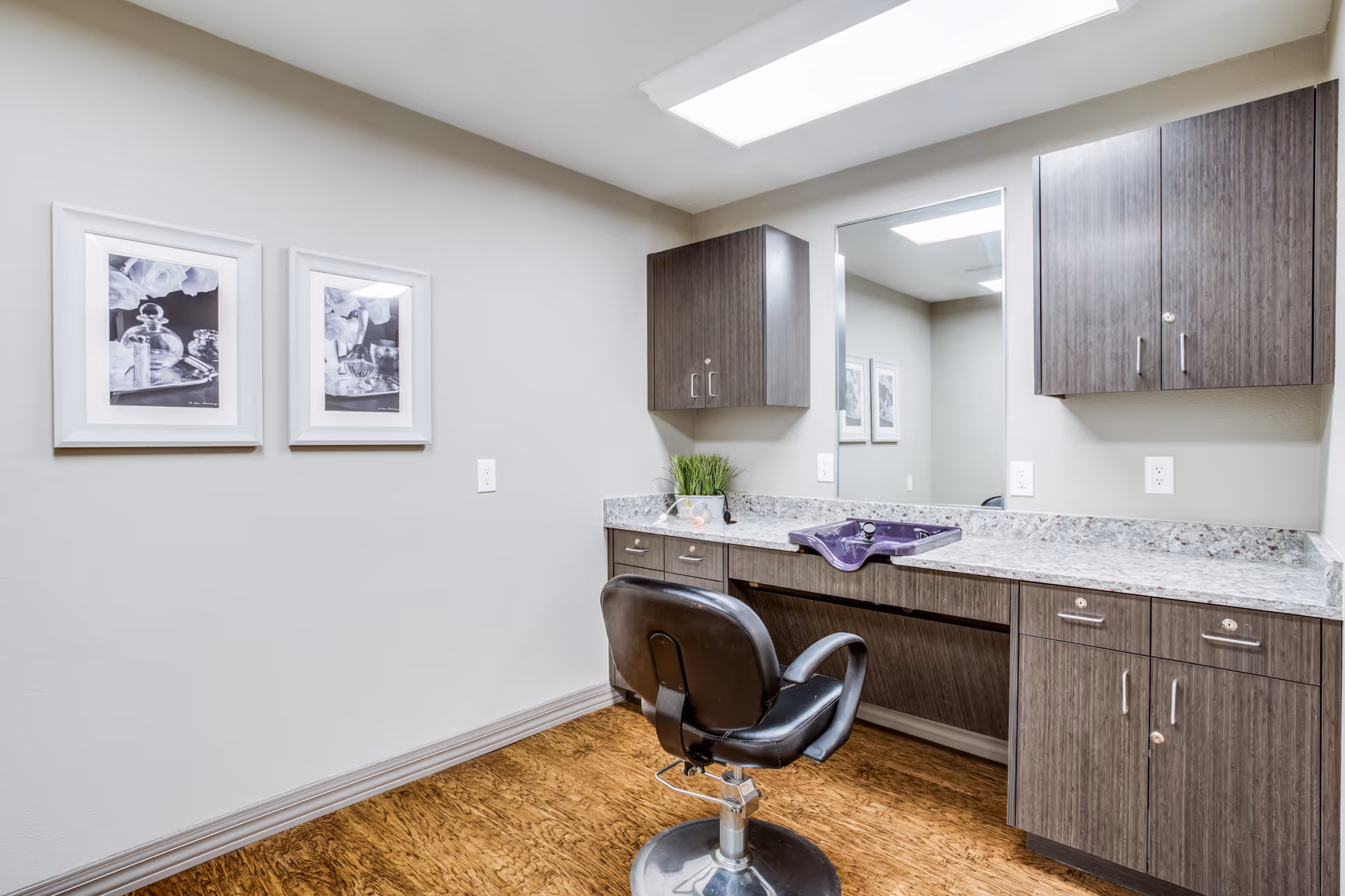 A small room with a black salon chair in front of a countertop with a purple sink basin. The countertop has a granite-like surface with dark wood cabinets above and below. A large mirror is mounted on the wall above the countertop. Two framed black and white pictures hang on the adjacent wall. The floor is a wood-like material and the walls are painted light gray.