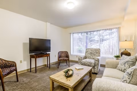 A cozy living room with a beige sofa, an armchair with patterned upholstery, two wooden chairs with patterned cushions, a wooden coffee table with decorative items and books, a flat-screen TV on a wooden stand, and a large window with vertical blinds letting in natural light.