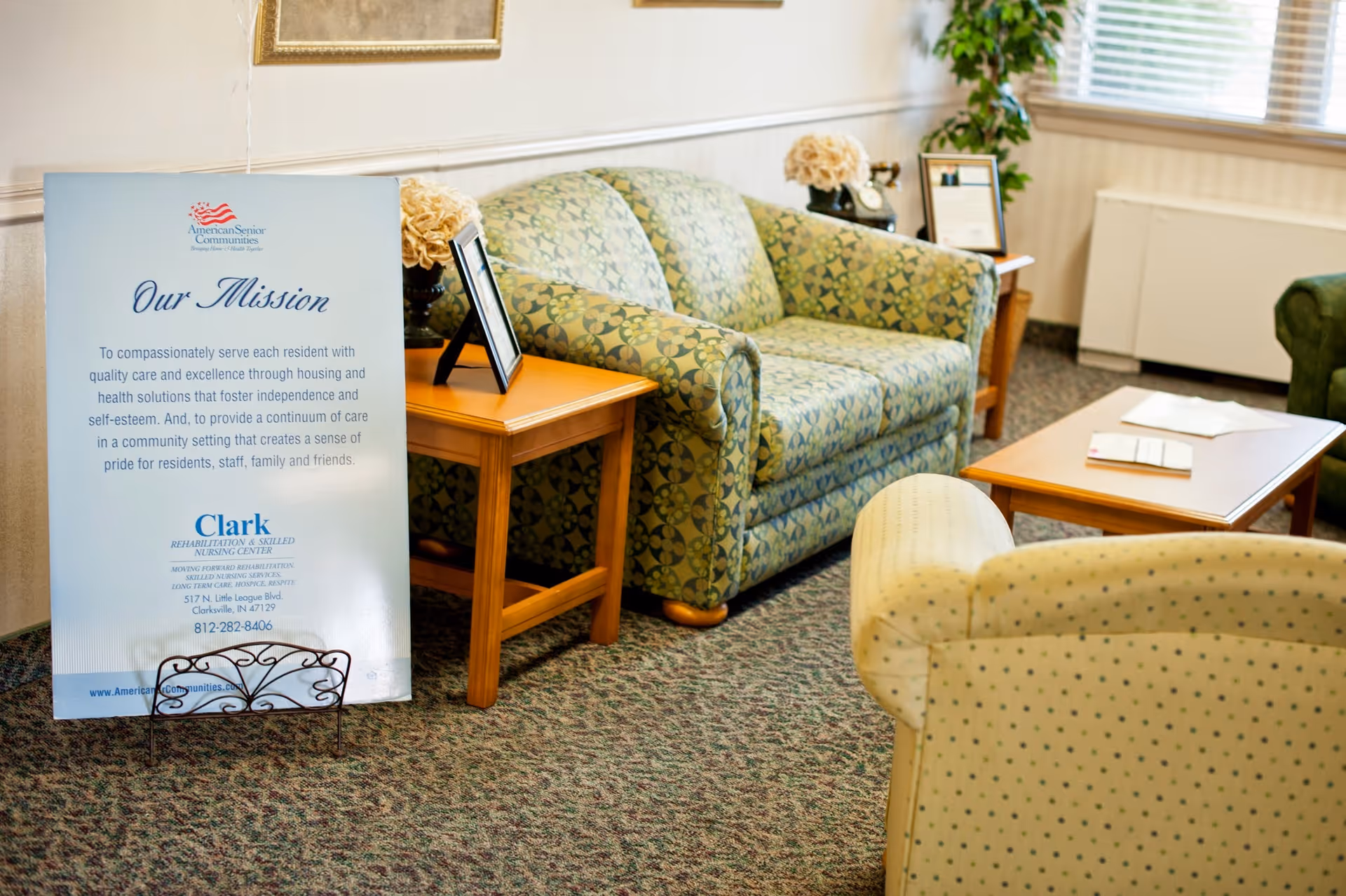 Seating area in a senior living facility with patterned sofas, armchairs, side tables and a large 'Our Mission' sign for Clark Rehabilitation and Skilled Nursing Center.