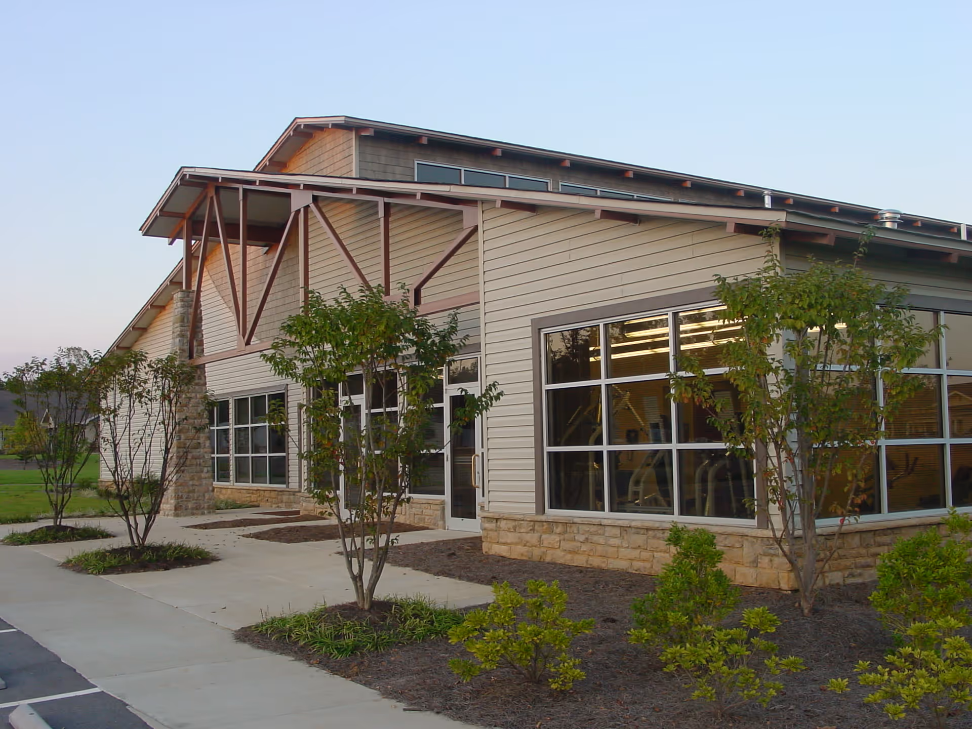 Exterior view of a modern single-story building with large windows, beige siding, stone accents, and a sloped roof. There are small trees and shrubs planted along the sidewalk in front of the building, and a parking lot is visible to the left.