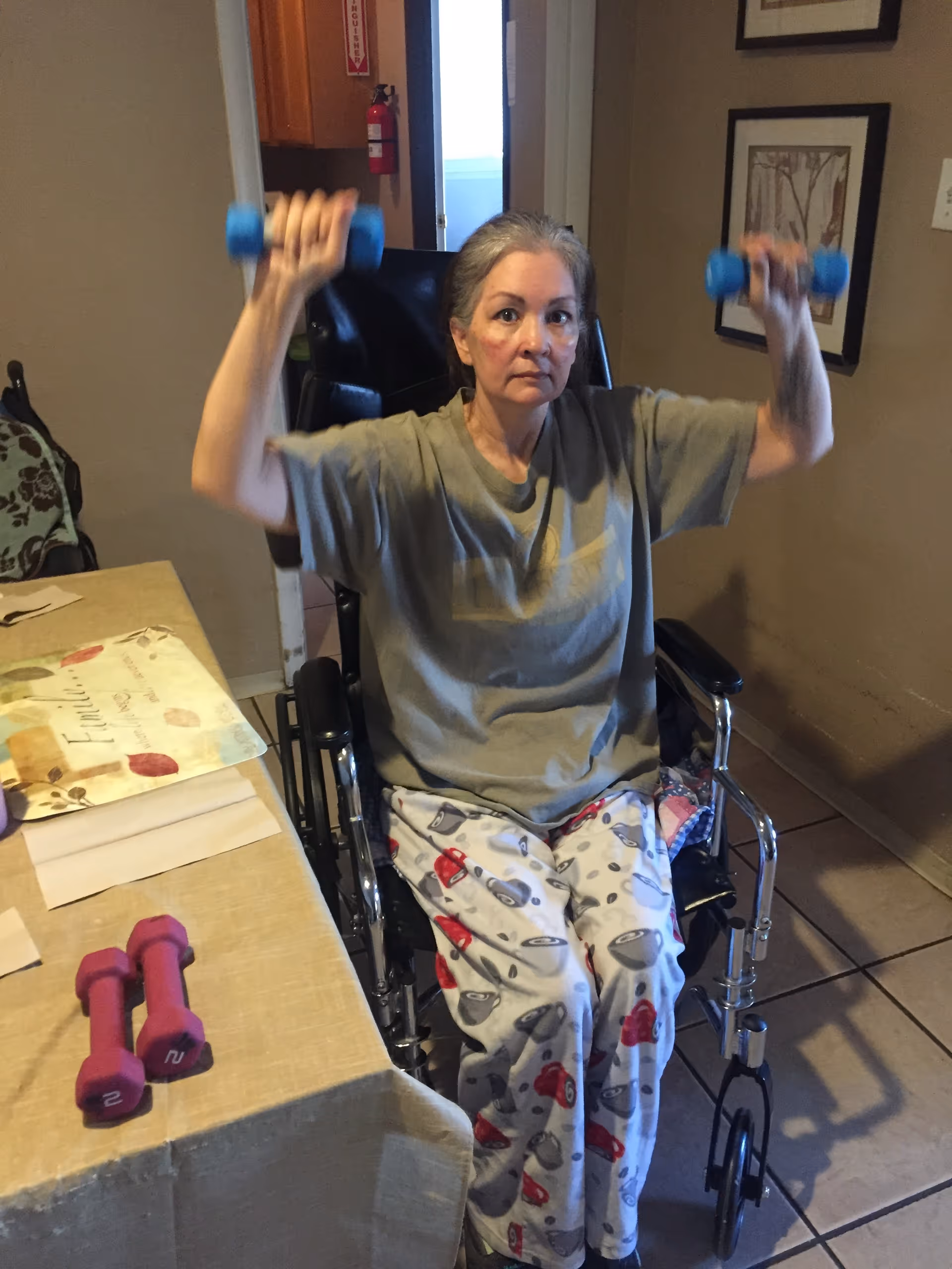 An elderly woman sitting in a wheelchair indoors, lifting blue dumbbells with both hands as part of an exercise routine. She is wearing a gray t-shirt and pajama pants with a pattern of kettlebells and weights. A table with a tablecloth and two pink dumbbells is visible to her left, and framed artwork hangs on the wall behind her.