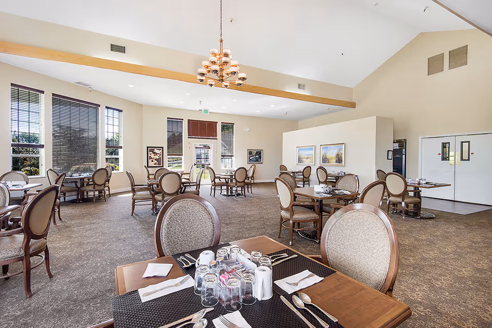 A spacious dining room with multiple tables and chairs arranged neatly. Each table is set with glasses, napkins, and silverware. Large windows with blinds allow natural light to fill the room. The ceiling is high with a wooden beam and a chandelier hanging in the center. The walls are decorated with framed artwork.