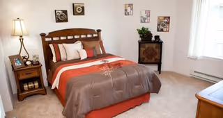 A cozy bedroom with a wooden bed frame and a bed made with brown, red, and beige bedding. There are several pillows on the bed. A wooden nightstand with a lamp, a photo frame, and a clock is beside the bed. The room has beige carpet, light-colored walls with framed artwork, a small cabinet with a plant and decorative items, and a window with sheer white curtains allowing natural light in.