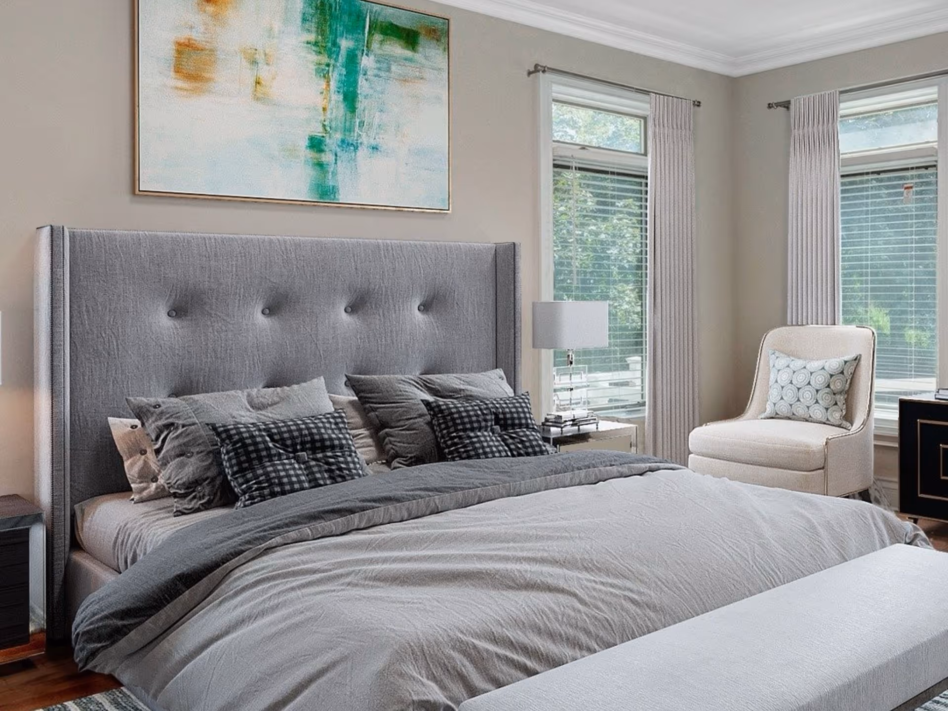 A modern bedroom featuring a large bed with a tall, tufted gray headboard and multiple pillows in shades of gray and black. There is a white cushioned chair with a decorative pillow near two windows with white blinds and curtains. A bedside table with a lamp is positioned next to the bed, and a large abstract painting with green and orange hues hangs above the headboard.