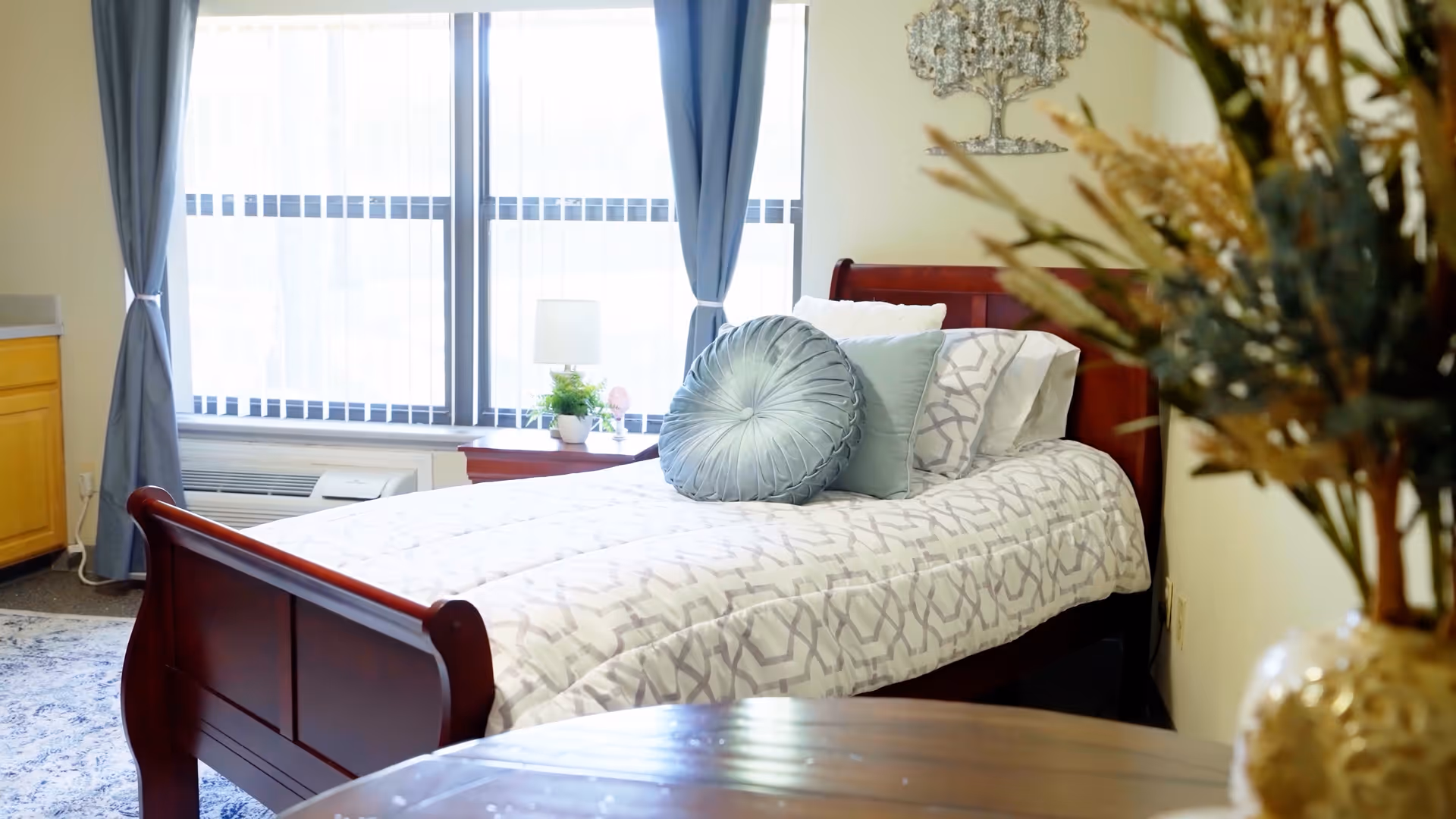 A bright and cozy bedroom in Forest Hills Assisted Living featuring a wooden bed with patterned bedding and decorative pillows. The room has large windows with sheer and blue curtains, a small bedside table with a lamp and plant, and a vase with dried flowers in the foreground.