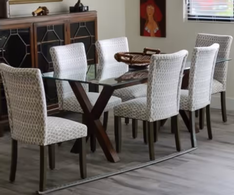 A modern dining room with a glass-top table supported by dark wooden legs and six upholstered chairs with a light geometric pattern. A wooden sideboard with decorative items is against the wall, and a colorful painting hangs above it. A window with blinds is visible on the right side.