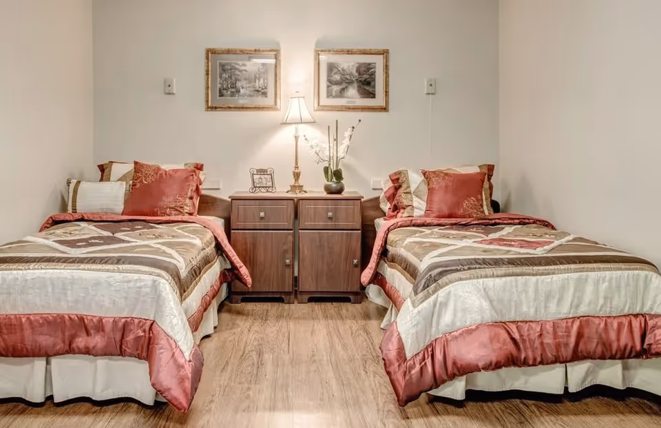 A bedroom with two twin beds separated by a wooden nightstand with two drawers. Each bed has a patterned bedspread in shades of beige, brown, and red, with multiple pillows. A table lamp and a small plant are on the nightstand. Two framed pictures hang on the wall above the nightstand. The floor is wooden.