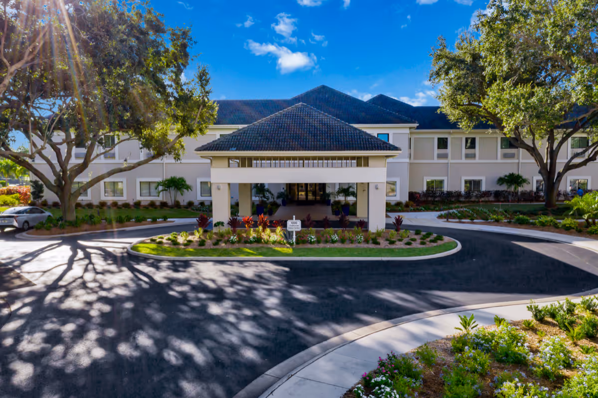 Front exterior view of a senior living facility named Arbor Trace with a covered entrance, surrounded by landscaped gardens, large trees, and a circular driveway under a bright blue sky with some clouds.