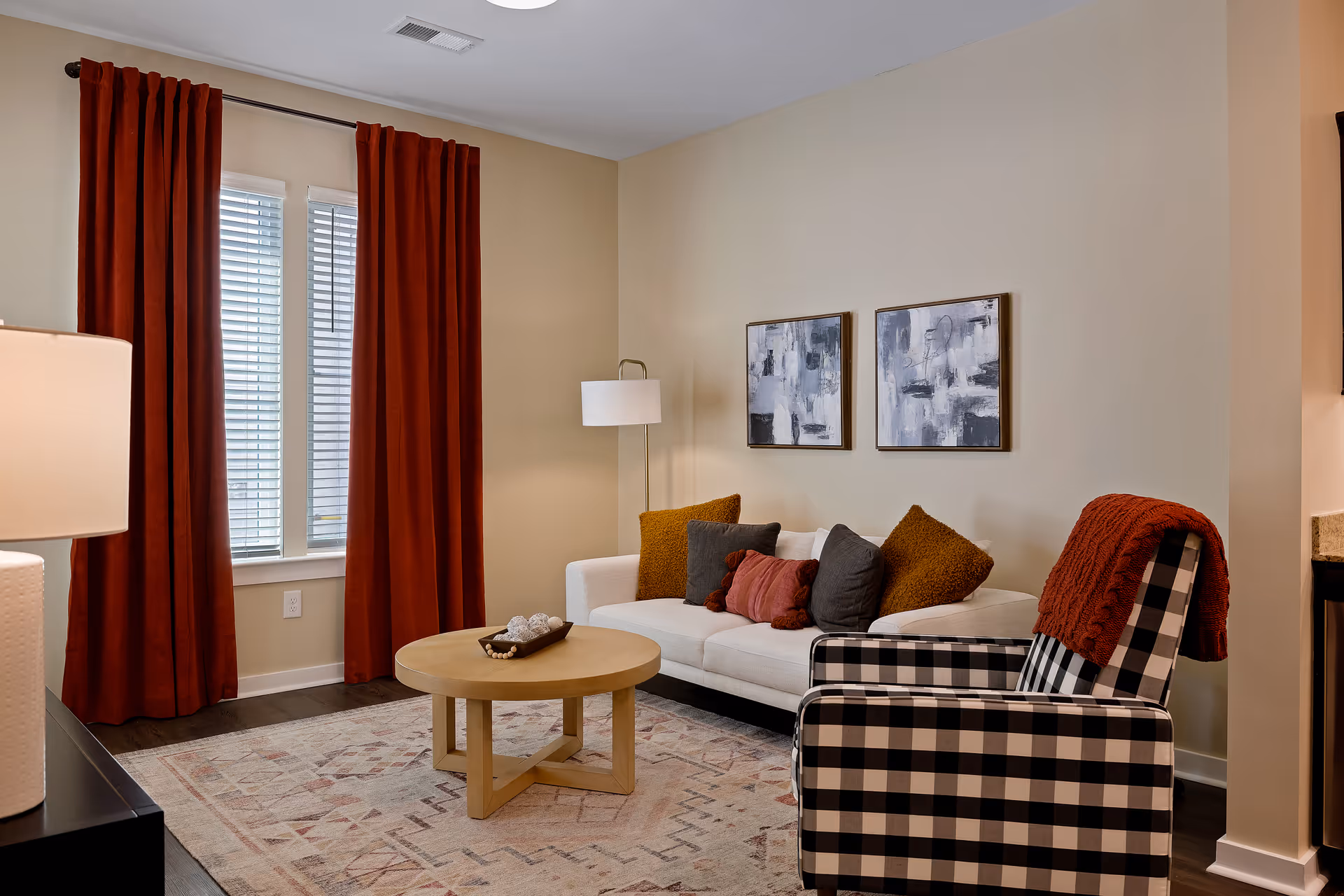 A cozy living room with a white sofa adorned with multiple cushions in brown, gray, and red tones. A round wooden coffee table sits in front of the sofa on a patterned area rug. To the right, there is a black and white checkered armchair with a red throw blanket draped over it. The room features beige walls with two abstract paintings hanging above the sofa, a floor lamp in the corner, and a window with white blinds and red curtains.