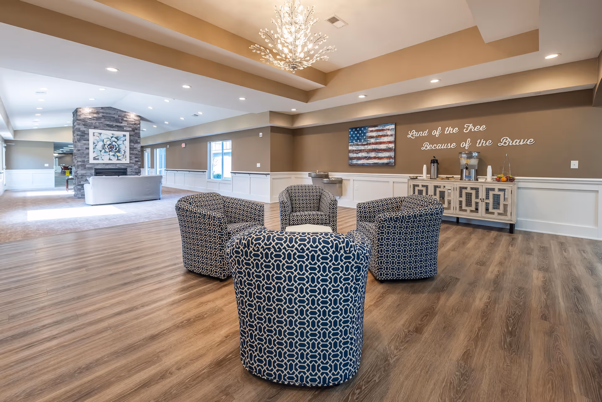 A spacious and well-lit common area in a senior living facility featuring four patterned armchairs arranged in a circle on wood flooring. In the background, there is a stone fireplace with a floral painting above it and a light gray sofa facing the fireplace. On the right wall, a decorative sideboard holds beverage dispensers and cups, with a painting of an American flag above it and the words 'Land of the Free Because of the Brave' displayed on the wall. The ceiling has recessed lighting and a decorative chandelier.