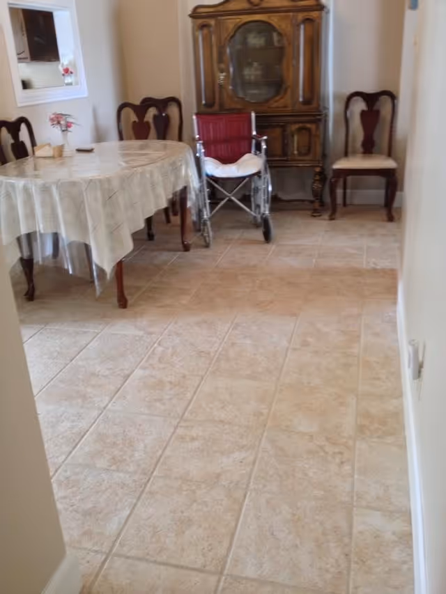 A dining area with a round table covered with a plastic tablecloth, surrounded by wooden chairs with cushioned seats. A red wheelchair is positioned near an antique wooden cabinet with glass doors. The floor is tiled with beige tiles.