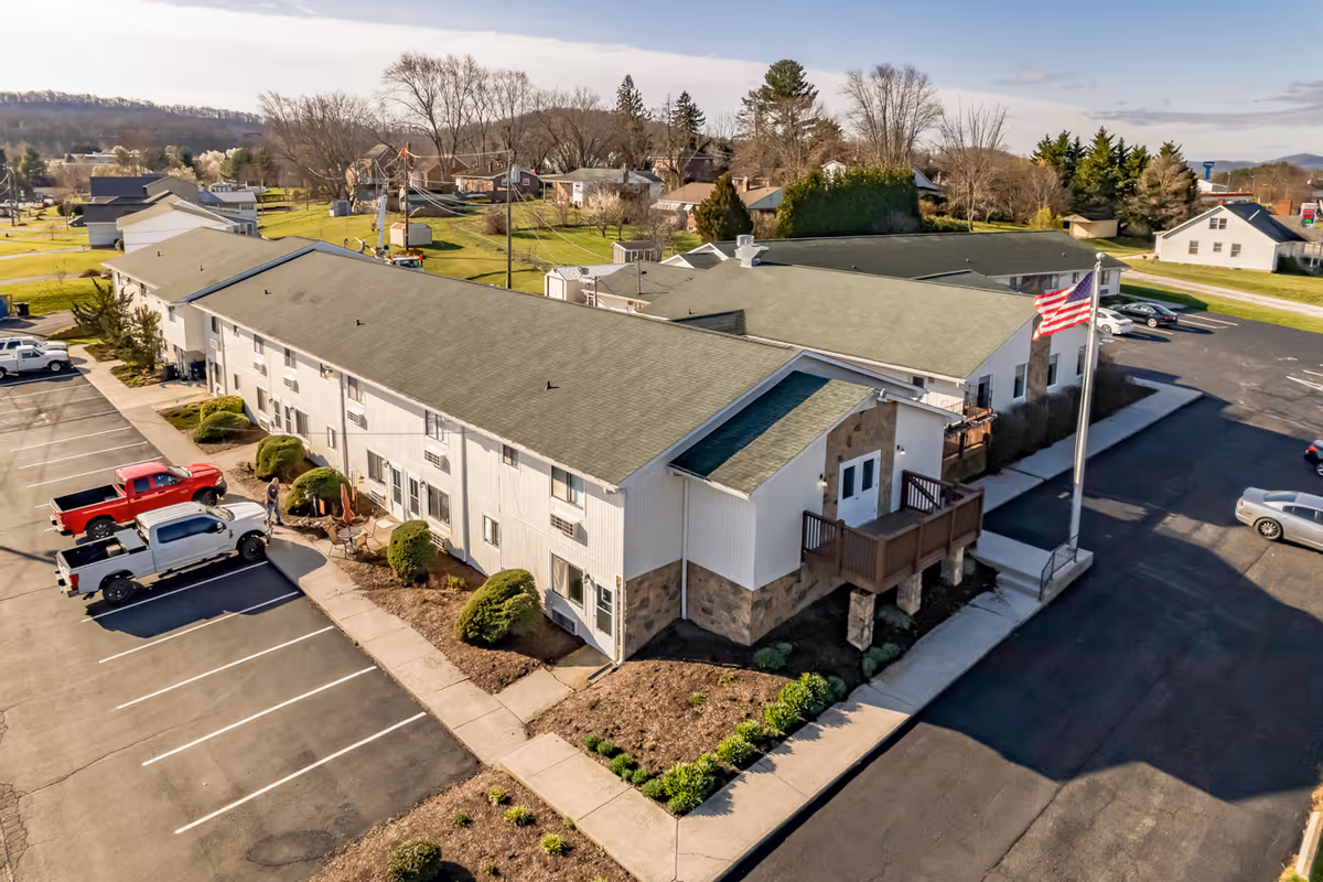 Aerial view of a two-story senior living facility building with a green roof and stone accents. The building is surrounded by a parking lot with several parked vehicles, landscaped bushes, and a sidewalk. An American flag is displayed on a flagpole near the entrance. The background shows a residential neighborhood with trees and houses under a partly cloudy sky.
