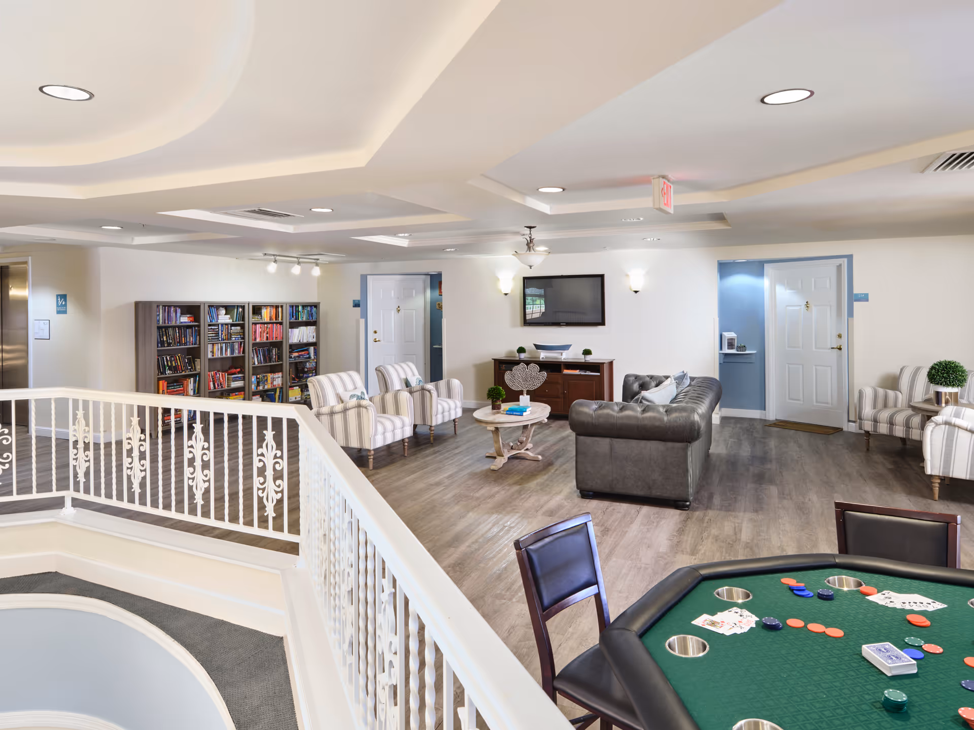 A spacious common area in a senior living facility featuring a poker table with playing cards and chips in the foreground, a seating area with a leather couch and striped armchairs, a coffee table, a TV mounted on the wall, and bookshelves filled with books. The room has light-colored walls, wood flooring, and recessed lighting in the ceiling.