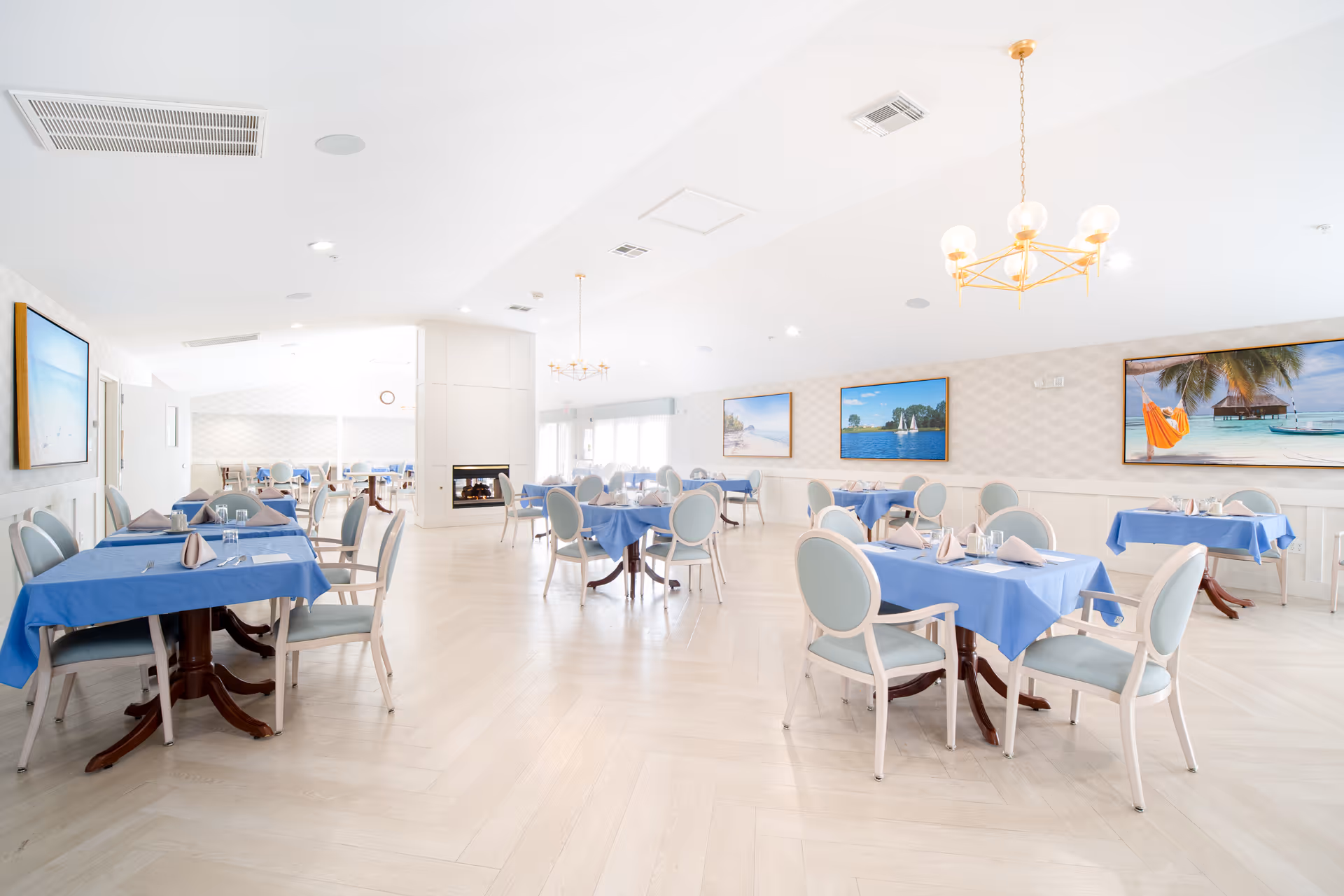 Bright and spacious dining room with multiple tables covered in blue tablecloths, each set with napkins and glassware. Light blue cushioned chairs surround the tables. The room features light-colored flooring, white walls with subtle patterns, and large framed pictures of beach and ocean scenes. Modern chandeliers hang from the ceiling, and a fireplace is visible in the background.