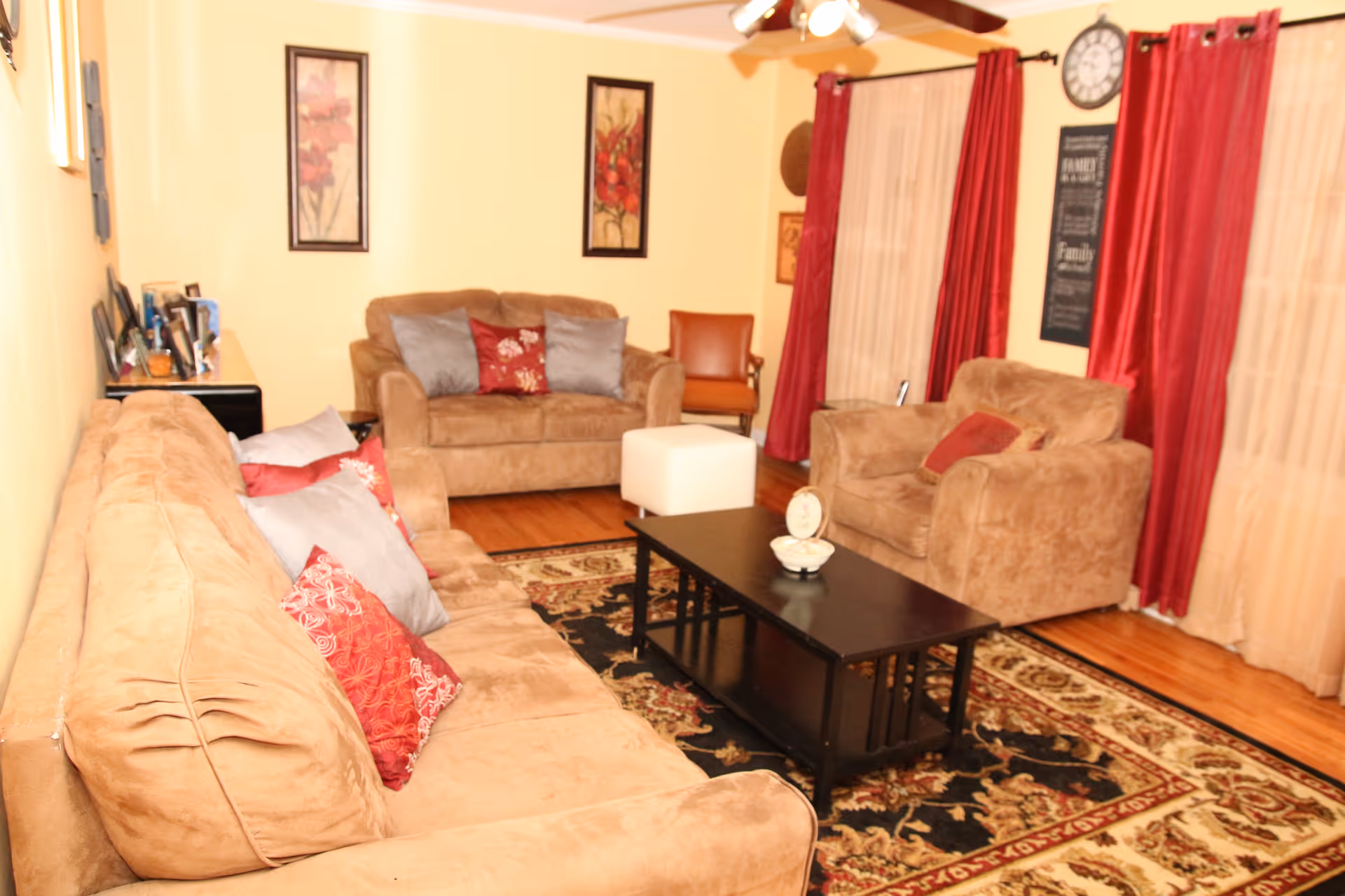 A cozy living room with beige sofas and armchairs adorned with red and gray pillows. A black coffee table sits on a patterned area rug in the center. The walls are painted light yellow and decorated with framed floral artwork and a clock. Red curtains hang over the windows, and a ceiling fan is visible above.