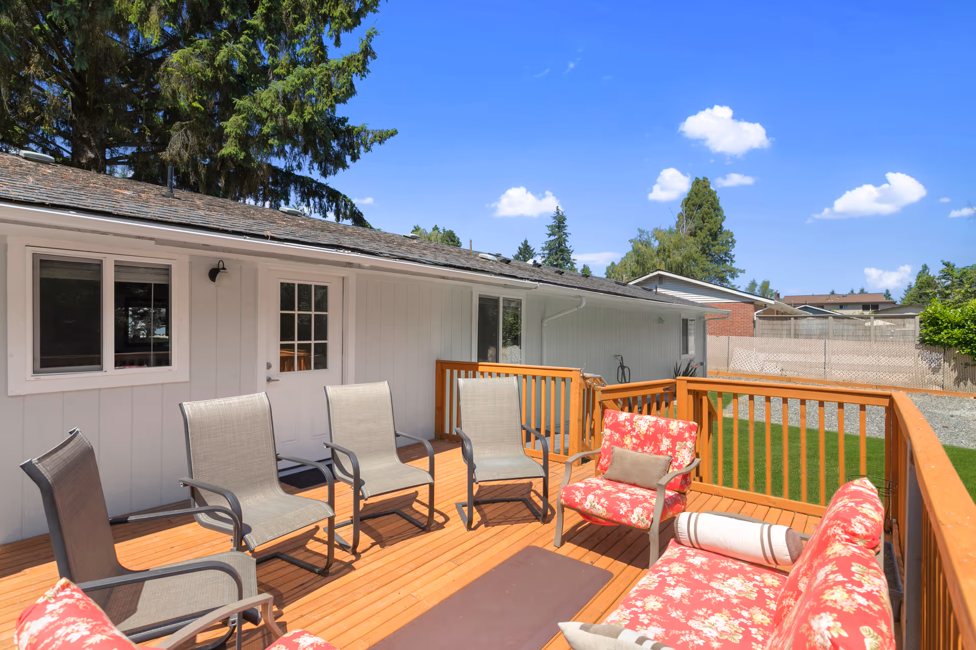 Outdoor wooden deck attached to a white building with several chairs and a cushioned sofa with floral patterns. The deck overlooks a green lawn and is surrounded by a wooden railing. Trees and a clear blue sky with a few clouds are visible in the background.