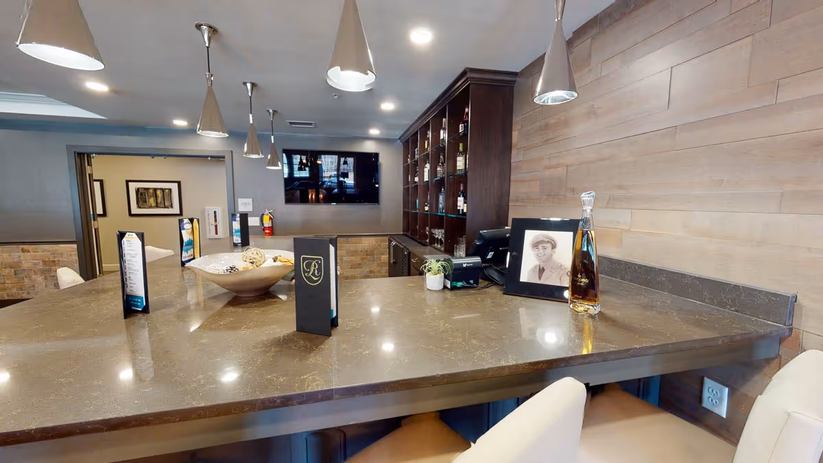 Interior view of a modern bar area with a polished stone countertop, several pendant lights hanging from the ceiling, a large wooden shelving unit stocked with bottles, a flat-screen TV mounted on the wall, and a few menus and decorative items on the counter.
