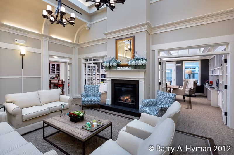A spacious and elegant living room with high ceilings, a fireplace in the center adorned with flower arrangements and a mirror above it. The room features comfortable white sofas and patterned armchairs arranged around a wooden coffee table with decorative items. The walls are painted in neutral tones with white trim, and there are adjoining rooms visible through open doorways, including a room with bookshelves and a desk.