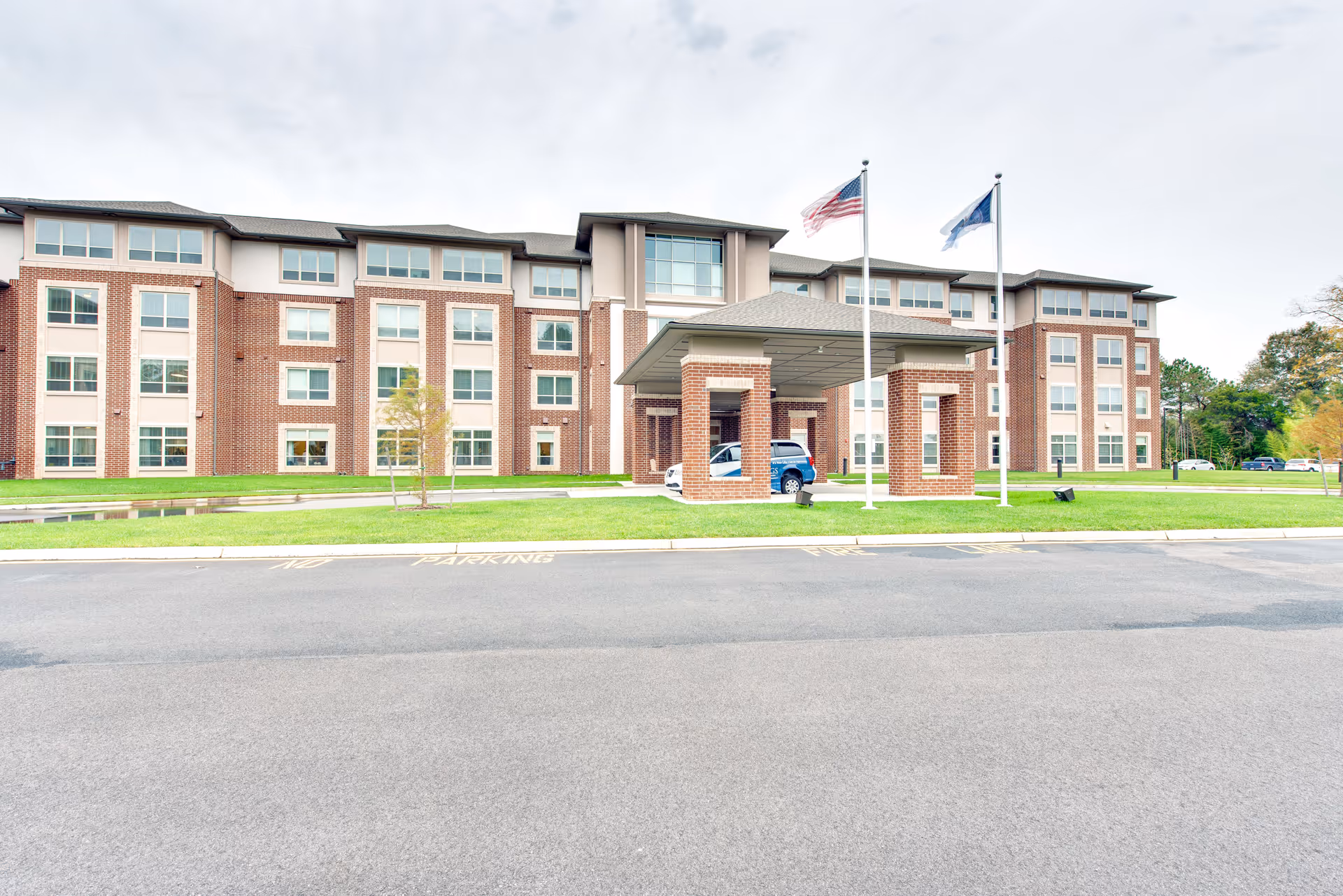 Front exterior view of a large brick senior living facility building with multiple windows, a covered entrance with brick pillars, two flagpoles with flags, and a driveway in front.