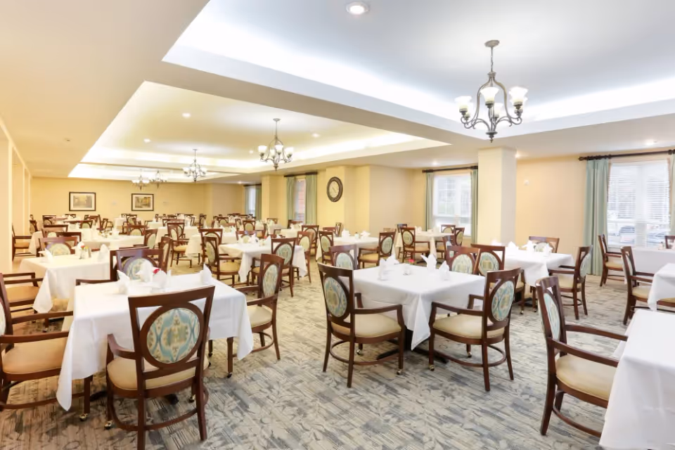 A spacious dining room with multiple tables covered in white tablecloths, each set with napkins and utensils. The room features wooden chairs with patterned upholstery, chandeliers hanging from the ceiling, large windows with curtains, and framed artwork on the walls.