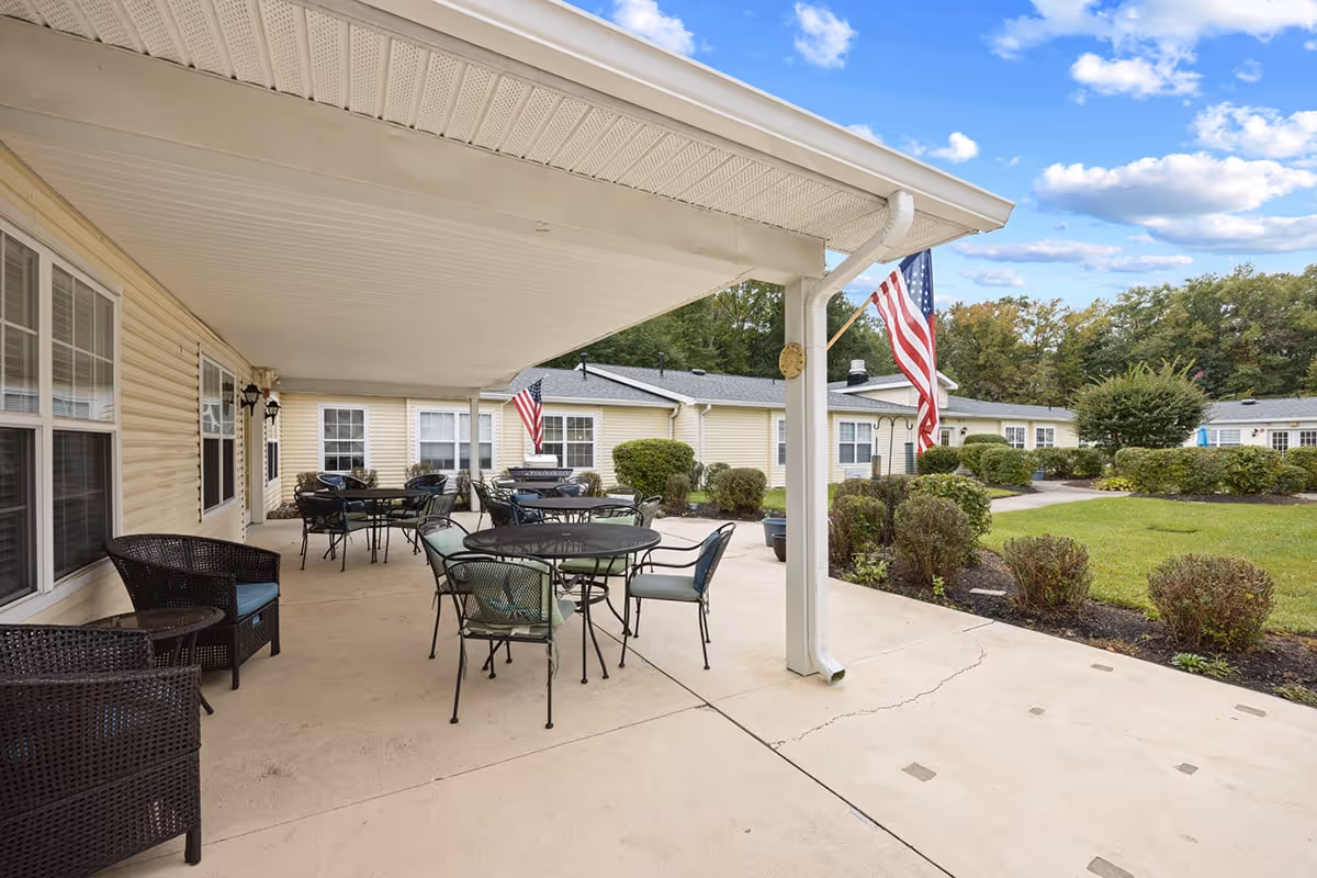 Covered outdoor patio area at Granville Place with several round tables and chairs arranged for seating. The patio overlooks a well-maintained garden with bushes and trees. American flags are displayed on the building and patio posts under a partly cloudy sky.