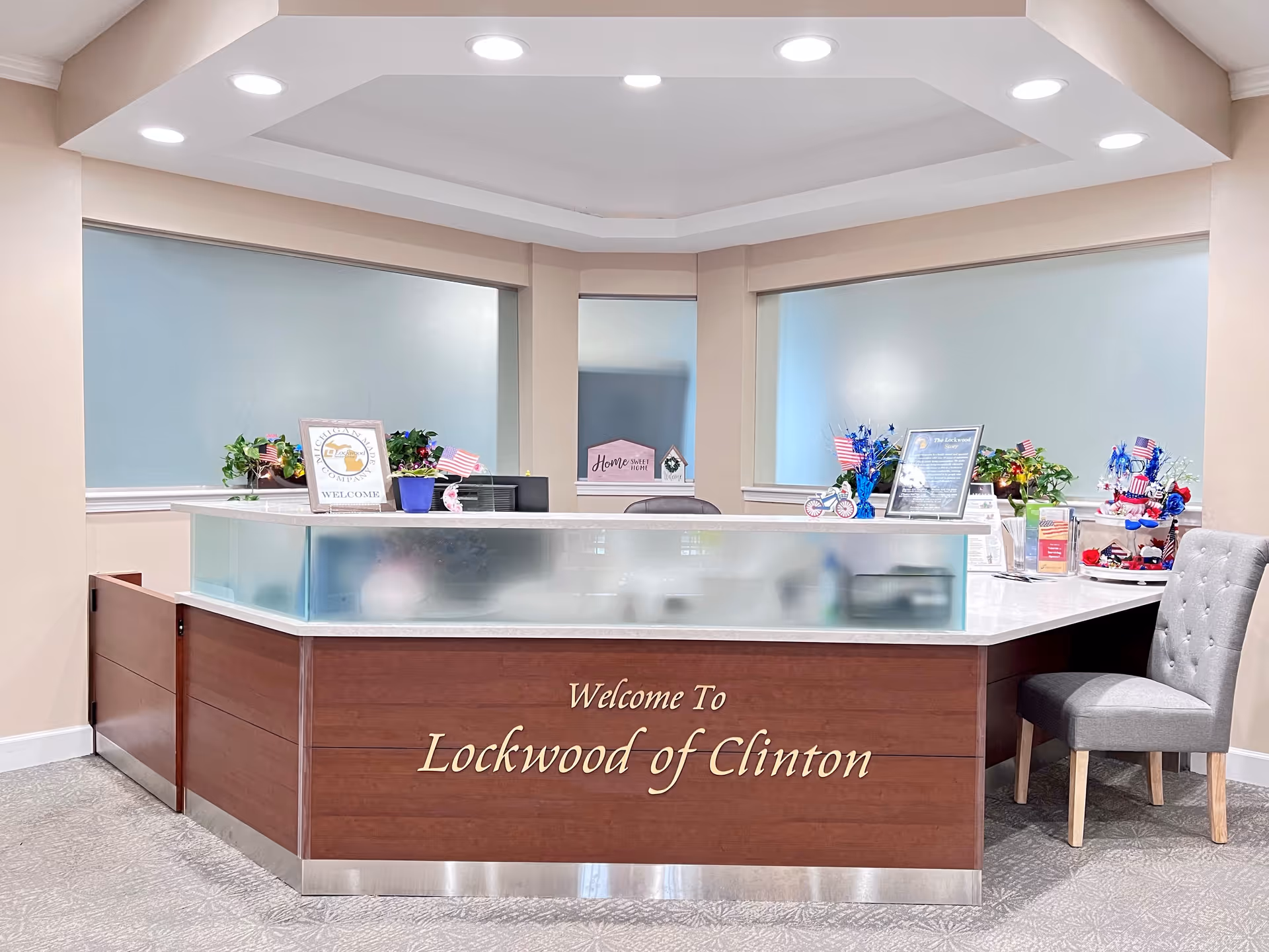 Reception desk area at Lockwood of Clinton with a wooden front panel displaying the facility name. The desk is decorated with small American flags, plants, and framed signs. There is a gray upholstered chair to the right and frosted glass panels on the desk. The walls are light beige with large windows covered by frosted glass.