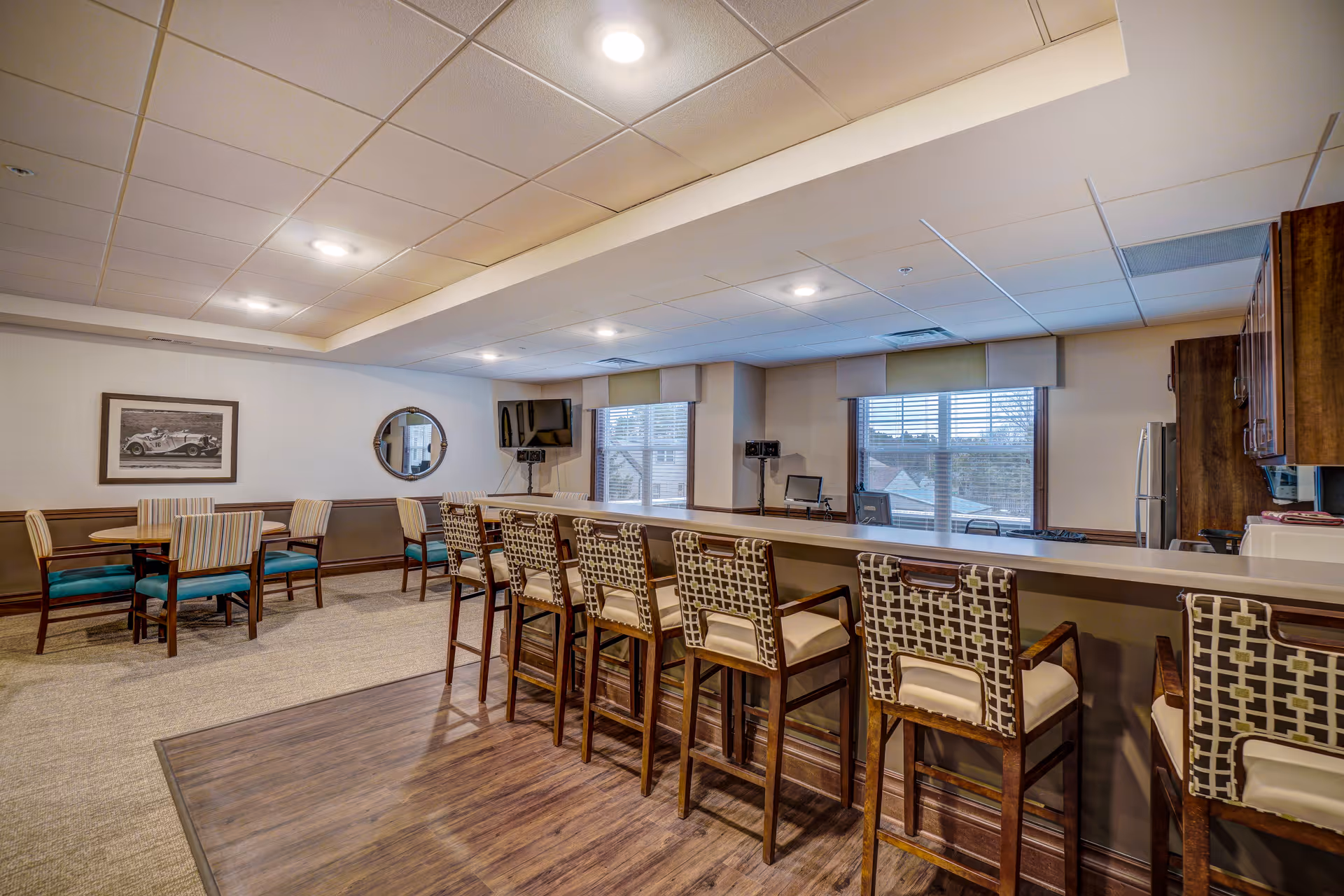 Interior view of a senior living facility common area with a long counter and six patterned cushioned bar stools. To the left, there are two round tables each surrounded by four chairs with striped upholstery. The room has large windows with blinds, a wall-mounted TV, a round mirror, and framed artwork. The flooring is a combination of carpet and wood, and the ceiling has recessed lighting.