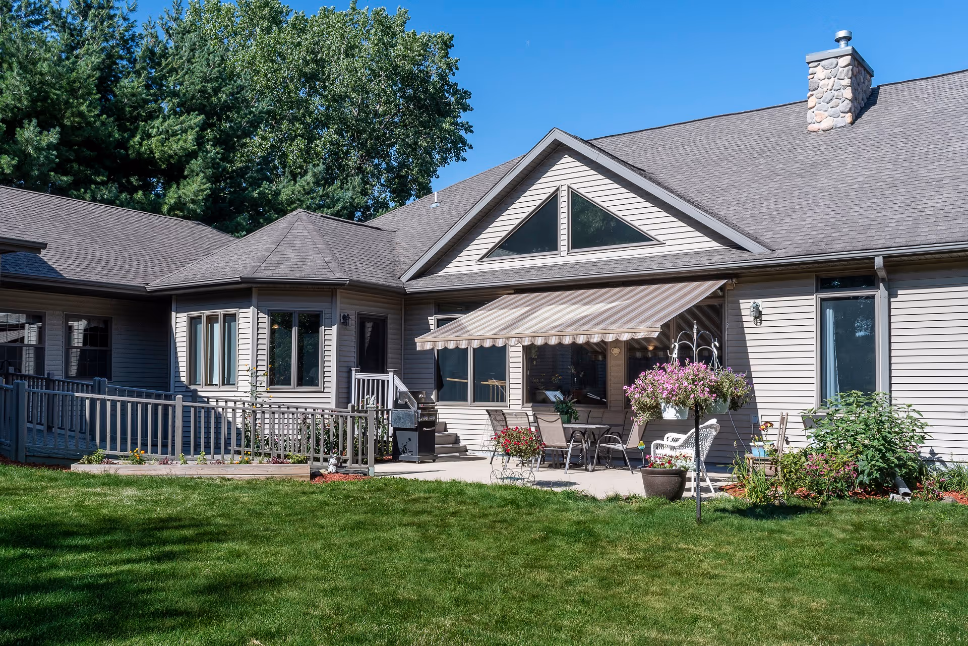 Exterior view of a single-story senior living facility building with beige siding, a stone chimney, and a patio area with outdoor furniture and potted plants. The patio has a striped awning, and there is a well-maintained green lawn in the foreground with trees in the background under a clear blue sky.