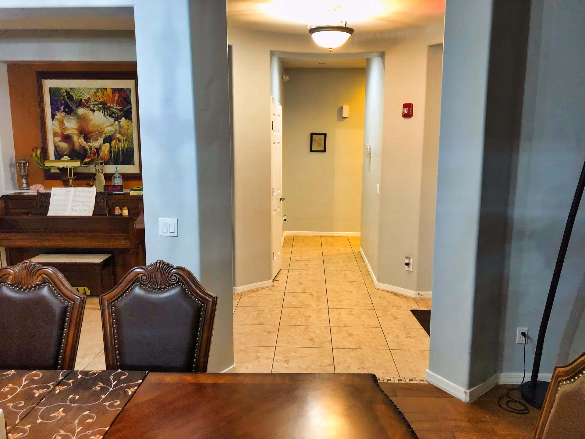 Interior view of a hallway with tiled floor leading to a door, seen from a dining area with a wooden table and two ornate chairs. To the left, there is a piano with sheet music and a colorful floral painting above it. The walls are painted light gray and the area is lit by a ceiling light fixture.
