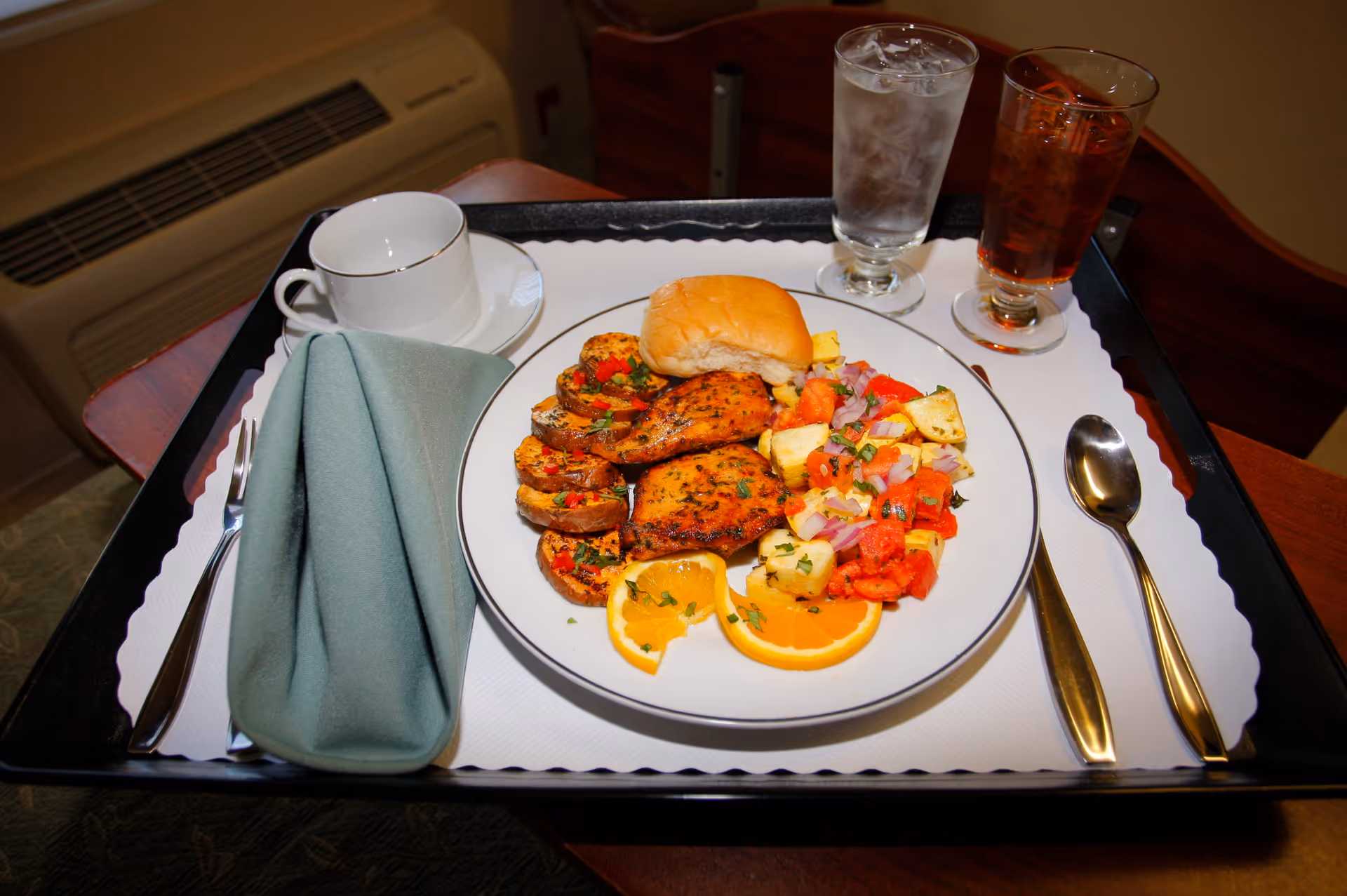 A plated meal of grilled chicken, roasted potatoes, vegetables, orange slices and a roll on a serving tray with utensils, a folded napkin, a cup, and two glasses.