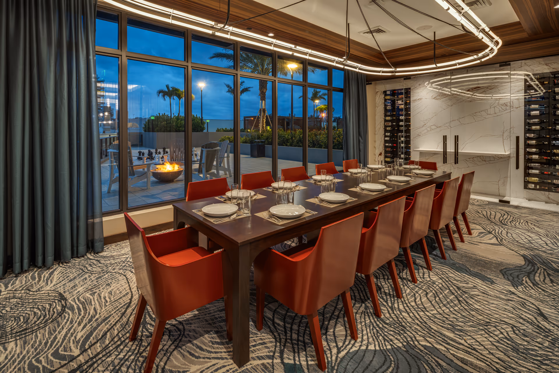A modern dining room with a long wooden table set for twelve people with white plates, glasses, and silverware. The room features red chairs, a patterned carpet, and large floor-to-ceiling windows showing an outdoor patio with chairs around a fire pit and palm trees in the background during dusk.