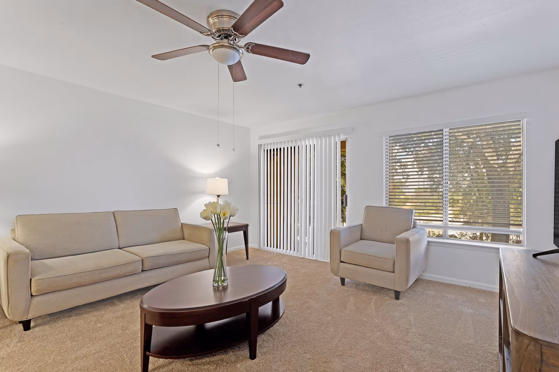 A bright living room with beige carpet, a beige sofa, a matching armchair, a dark wooden oval coffee table with a vase of flowers, a side table with a lamp, a ceiling fan, and large windows with blinds and vertical sliding glass door.