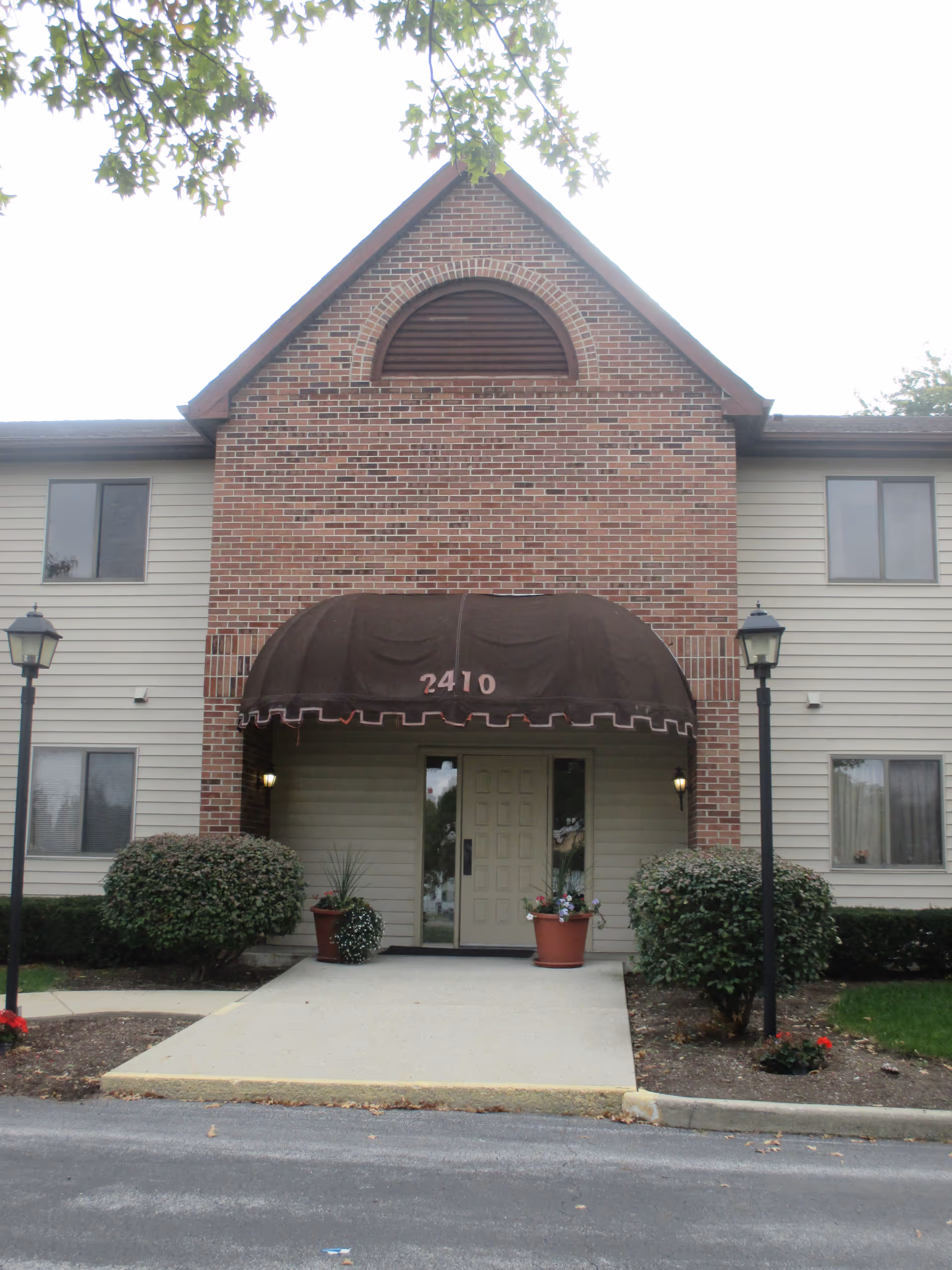 Entrance of a two-story brick-and-siding apartment building with a brown awning reading "2410", flanked by lamp posts and potted plants.