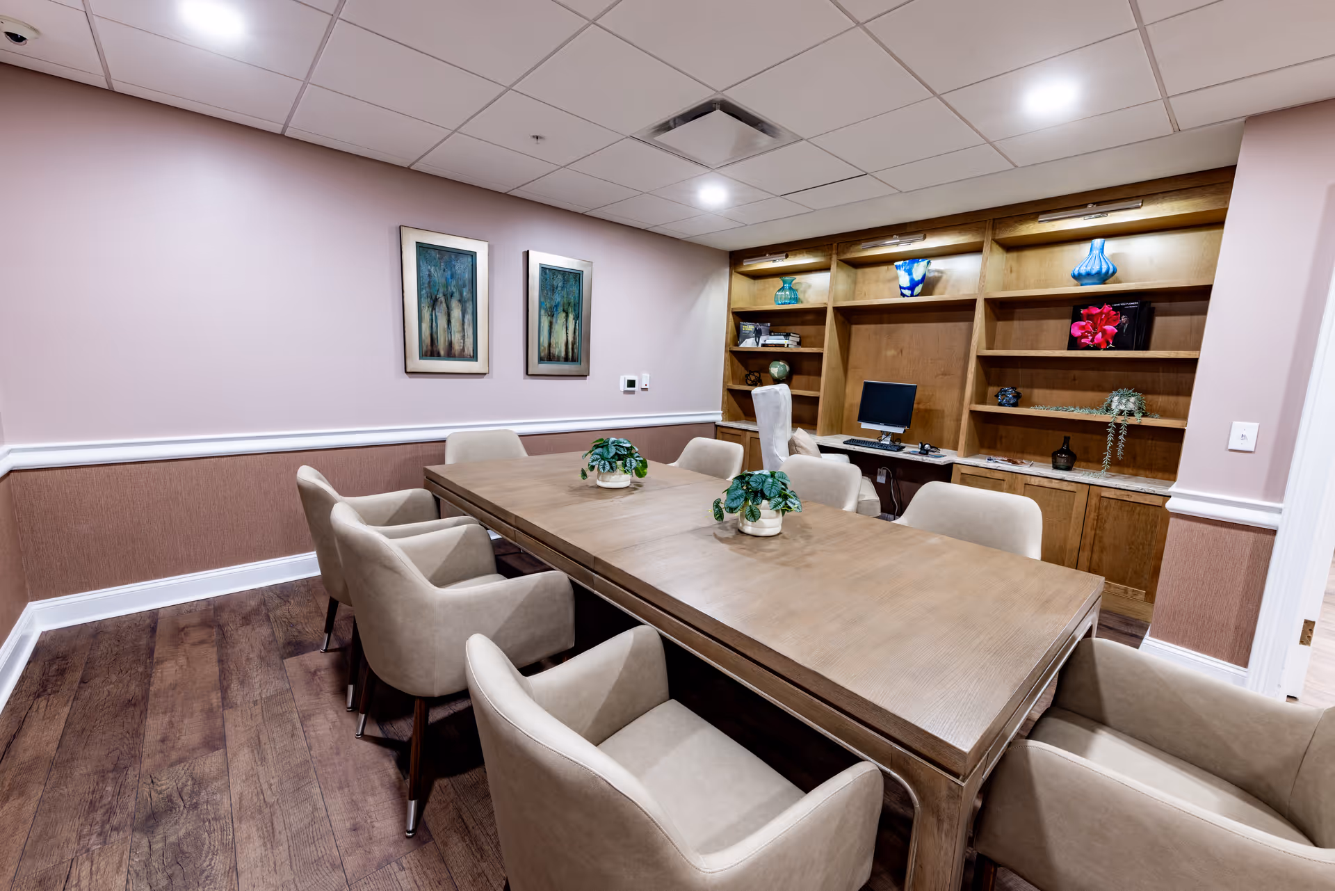 A conference or meeting room with a large wooden table surrounded by beige upholstered chairs. The room has wooden flooring, light pink walls with white trim, and two framed paintings on one wall. A built-in wooden shelving unit with decorative items and a computer is visible at the far end of the room.