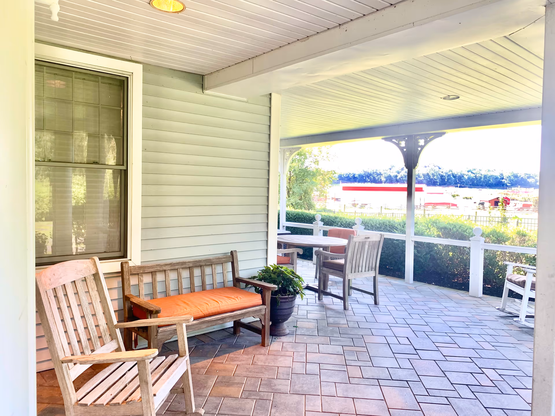 Covered outdoor patio area with wooden chairs and a bench with an orange cushion, a potted plant, and a view of greenery and buildings in the distance.