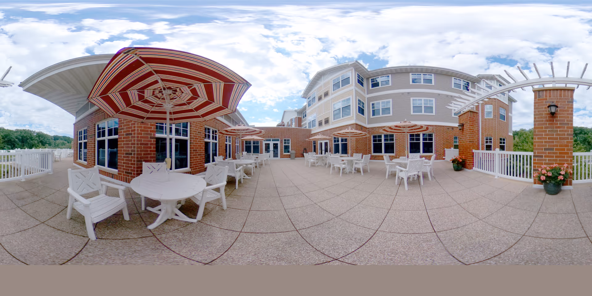Outdoor patio area at Wittenberg Village: Lutheran Life Communities featuring multiple white tables and chairs with red and white striped umbrellas. The patio is paved and surrounded by a white railing, with brick pillars and a multi-story building in the background under a partly cloudy sky.
