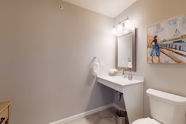 A small bathroom with a white toilet, a wall-mounted sink with a mirror above it, a towel ring holding a white towel, a small trash can underneath the sink, and a colorful painting on the wall depicting a person in a blue coat near a waterfront with the Eiffel Tower in the background.