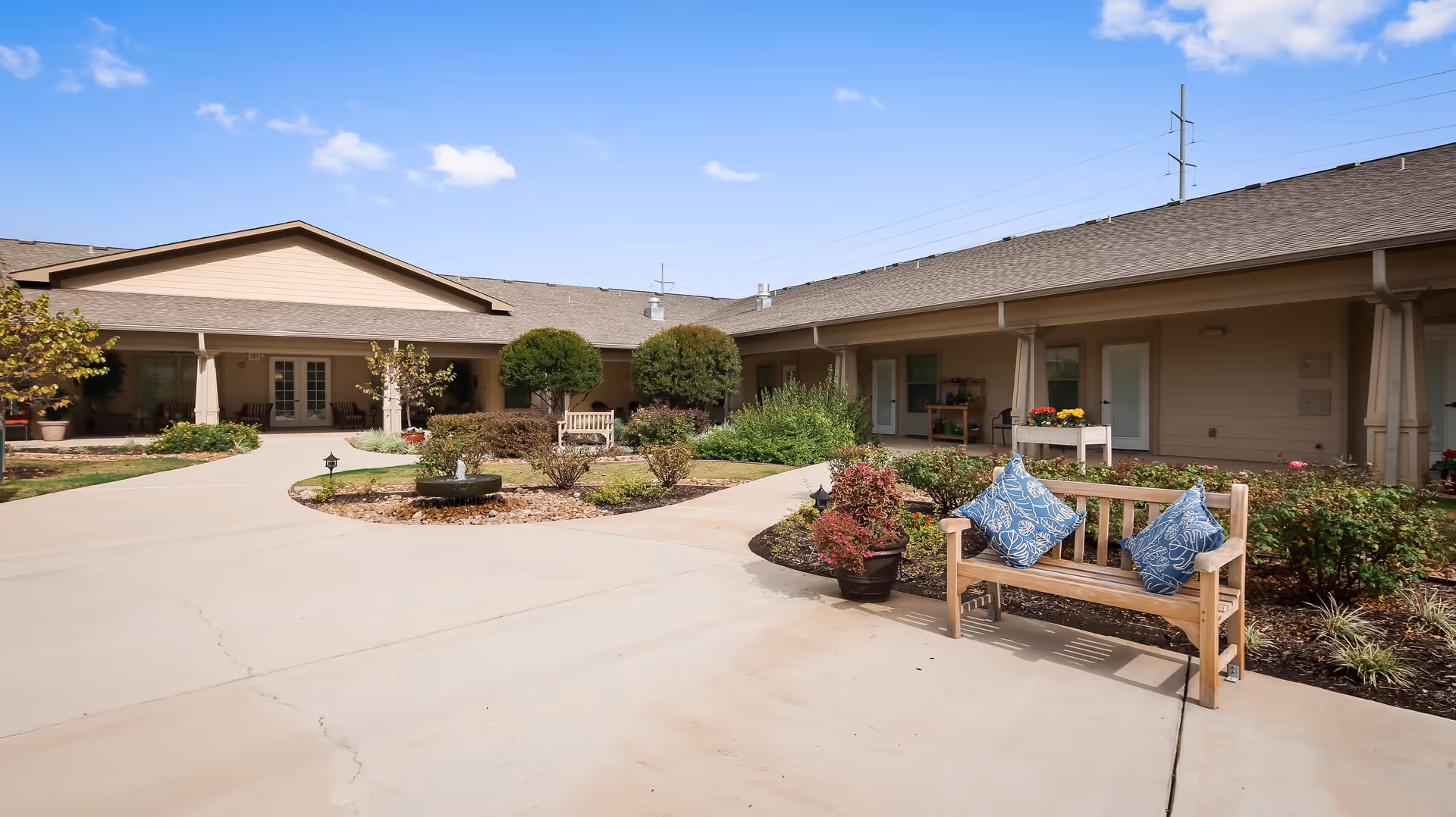 Outdoor courtyard area of Meadowood Assisted Living and Memory Care featuring a paved walkway, landscaped garden beds with shrubs and small trees, wooden benches with blue patterned cushions, and a beige building with multiple doors and windows under a clear blue sky.