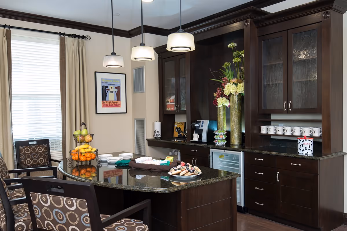 Communal kitchen area with a central island, dark wood cabinets, pendant lights, and fruit and pastries on the counter.