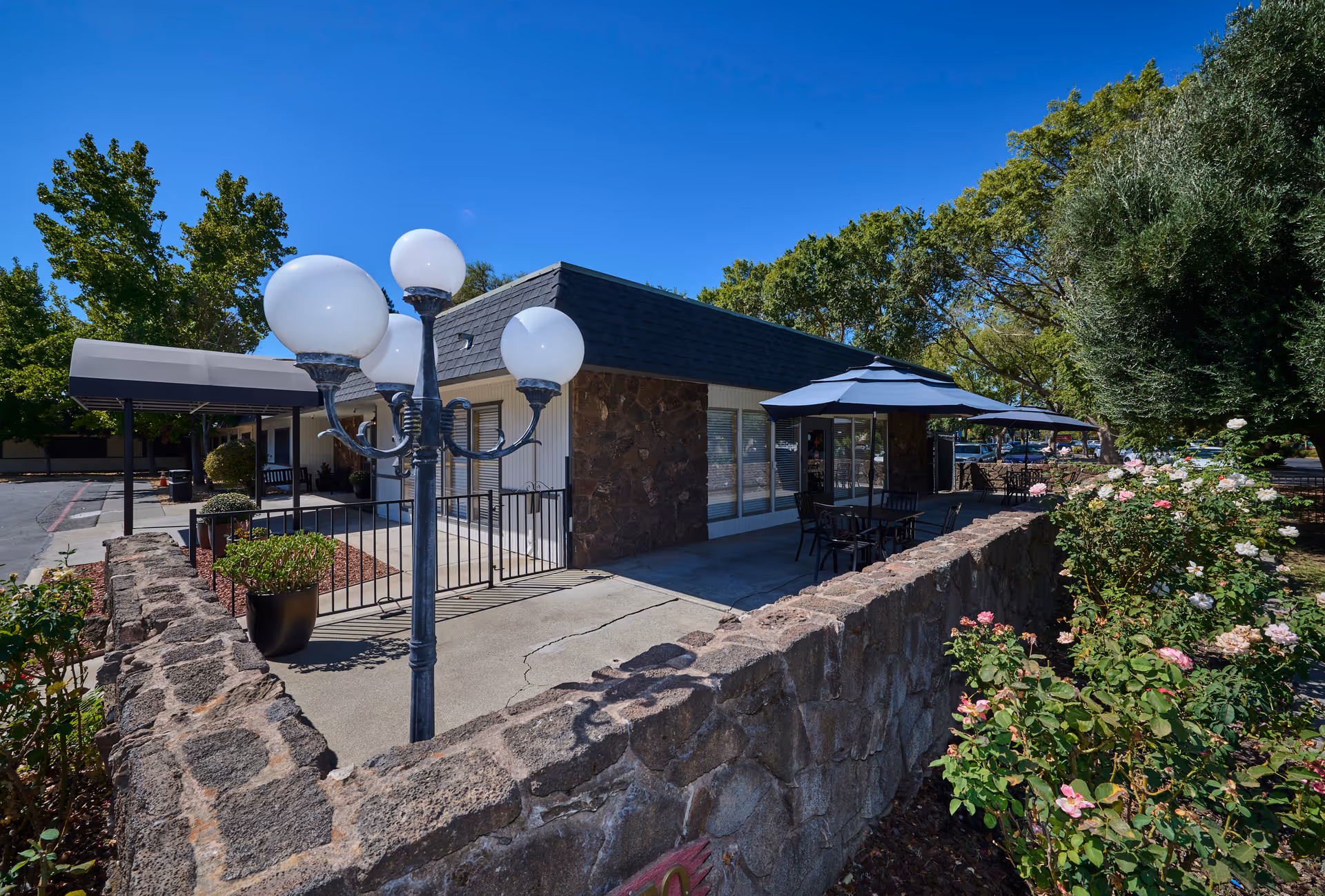 Outdoor patio area of Courtyard Healthcare Center with stone walls, a black metal lamp post with round white lights, tables with umbrellas, and surrounding greenery including rose bushes and trees under a clear blue sky.
