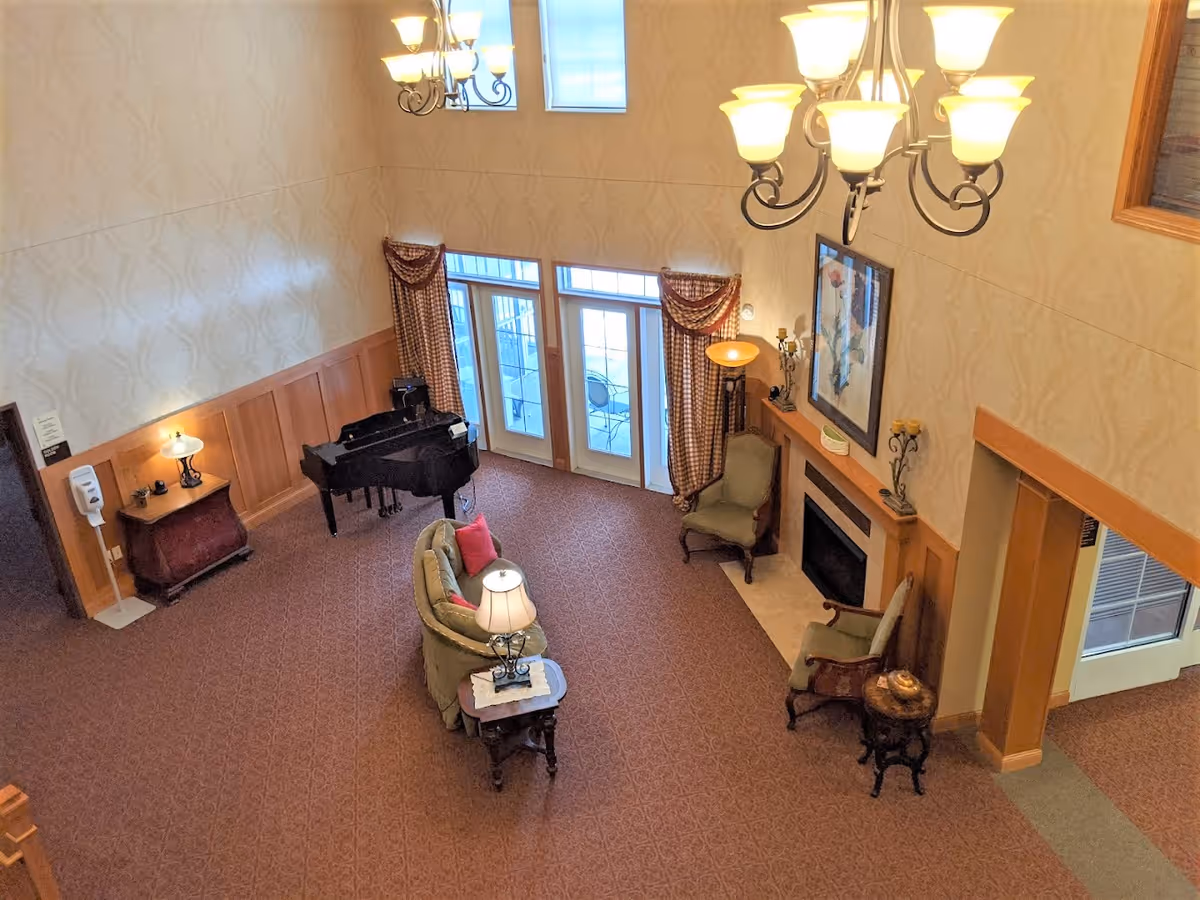 A high-ceilinged common lounge with seating, a grand piano, fireplace, and chandelier viewed from above.