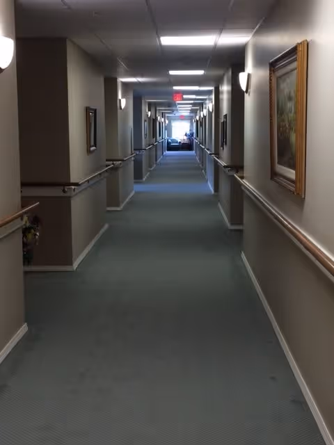 A long, carpeted hallway in an assisted living facility with handrails on both sides, wall-mounted lights, framed paintings, and an exit sign at the far end near a seating area.