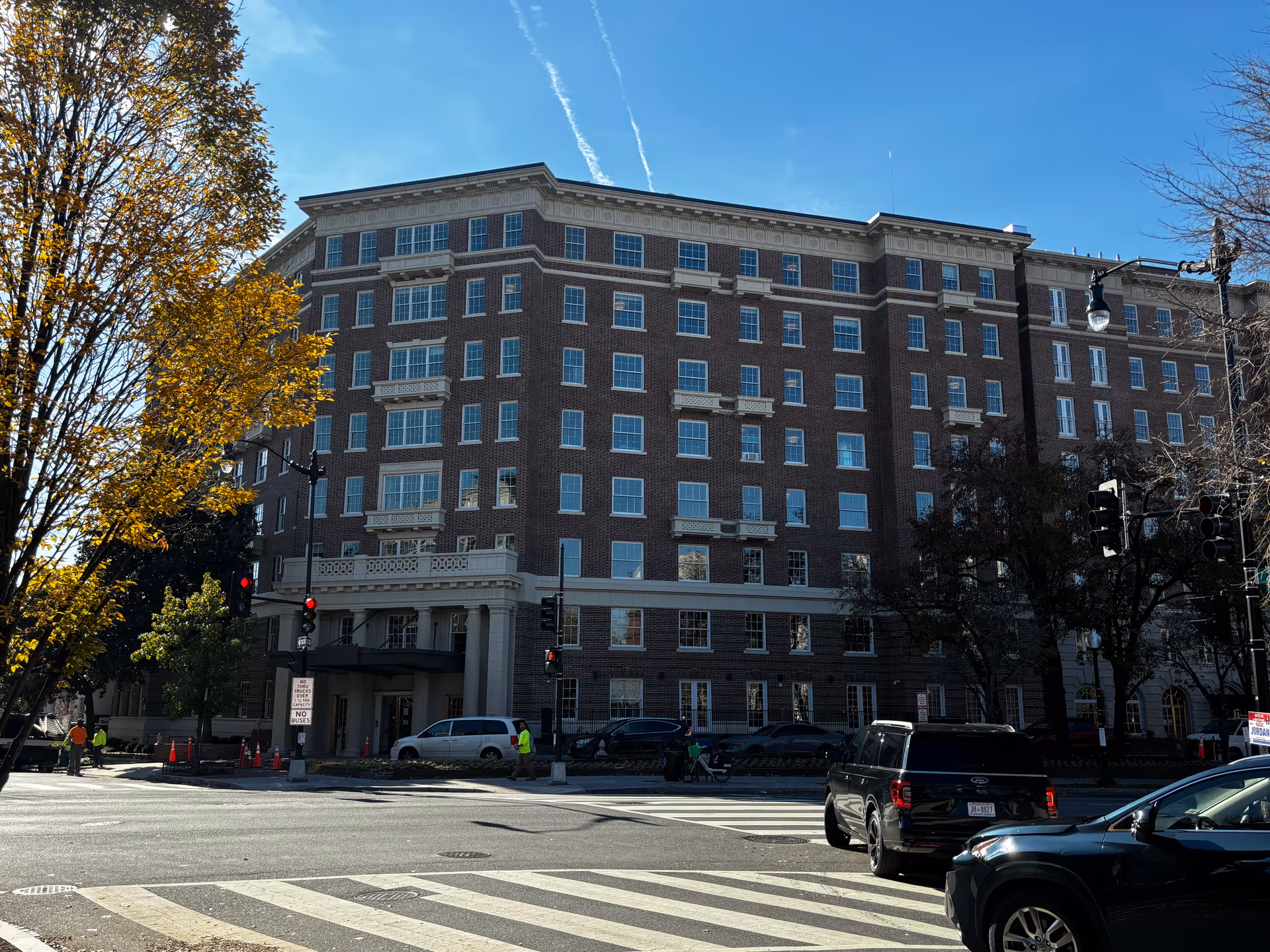 Large brick multi-story building at a street intersection with cars, a crosswalk, and trees in the foreground.