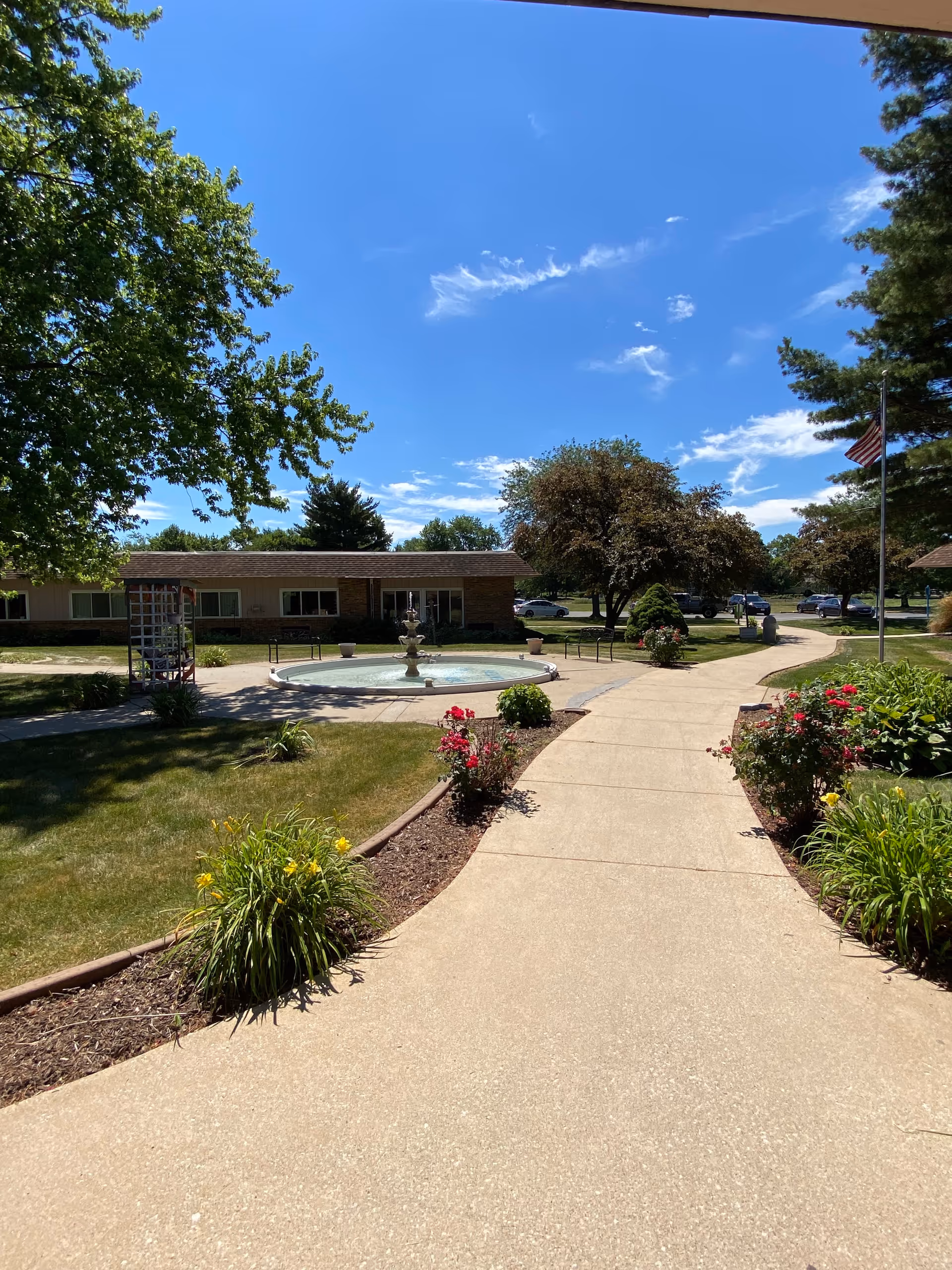 A sunny outdoor pathway leading to a circular fountain surrounded by greenery and flowers. Trees and bushes are visible along the path, with a building in the background and a clear blue sky overhead.