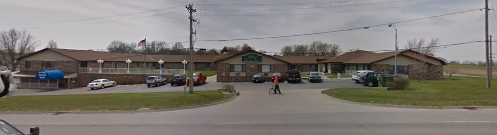 Exterior view of The Village Retirement Community building with a parking lot in front. Several cars are parked, and two people are walking near the entrance. The building is single-story with a stone facade and a sign displaying the facility's name.