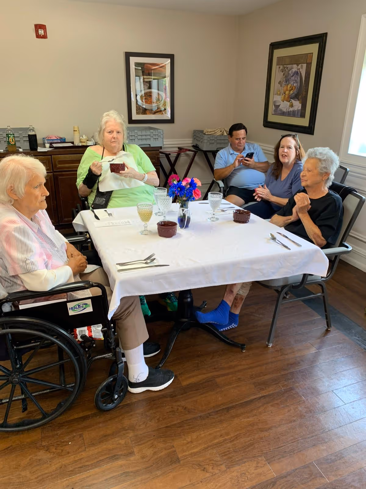 Five elderly people sitting around a dining table in a room with wooden floors and light-colored walls. The table is covered with a white tablecloth and has glasses, bowls, and a small vase with colorful flowers. Two framed pictures hang on the walls, and a window lets in natural light. One person is in a wheelchair, and another is using a smartphone.