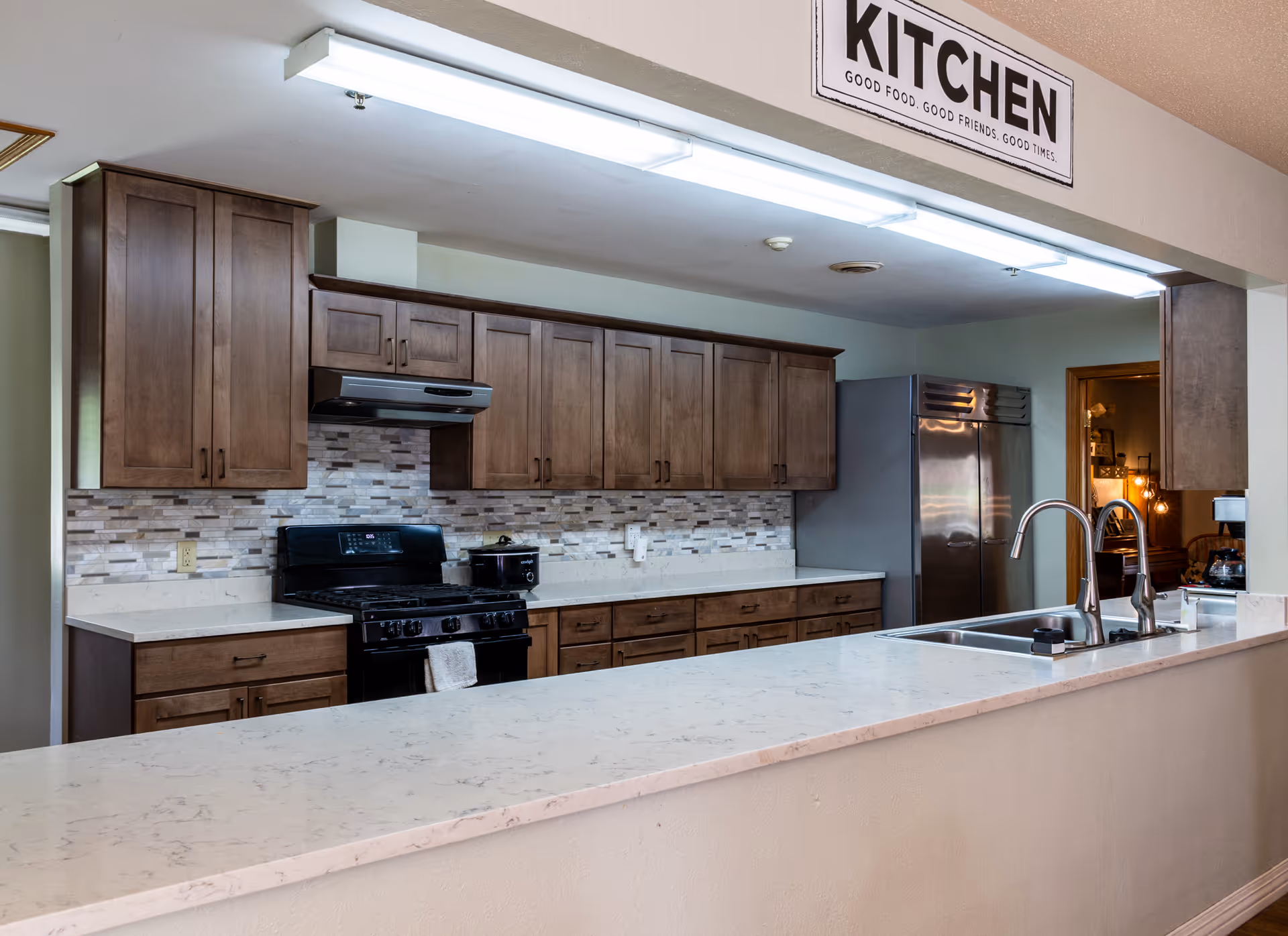 A modern kitchen with wooden cabinets, a black stove with a hood, a stainless steel refrigerator, and a long white marble countertop with a double sink and two faucets. Above the kitchen area is a sign that reads 'KITCHEN GOOD FOOD. GOOD FRIENDS. GOOD TIMES.'