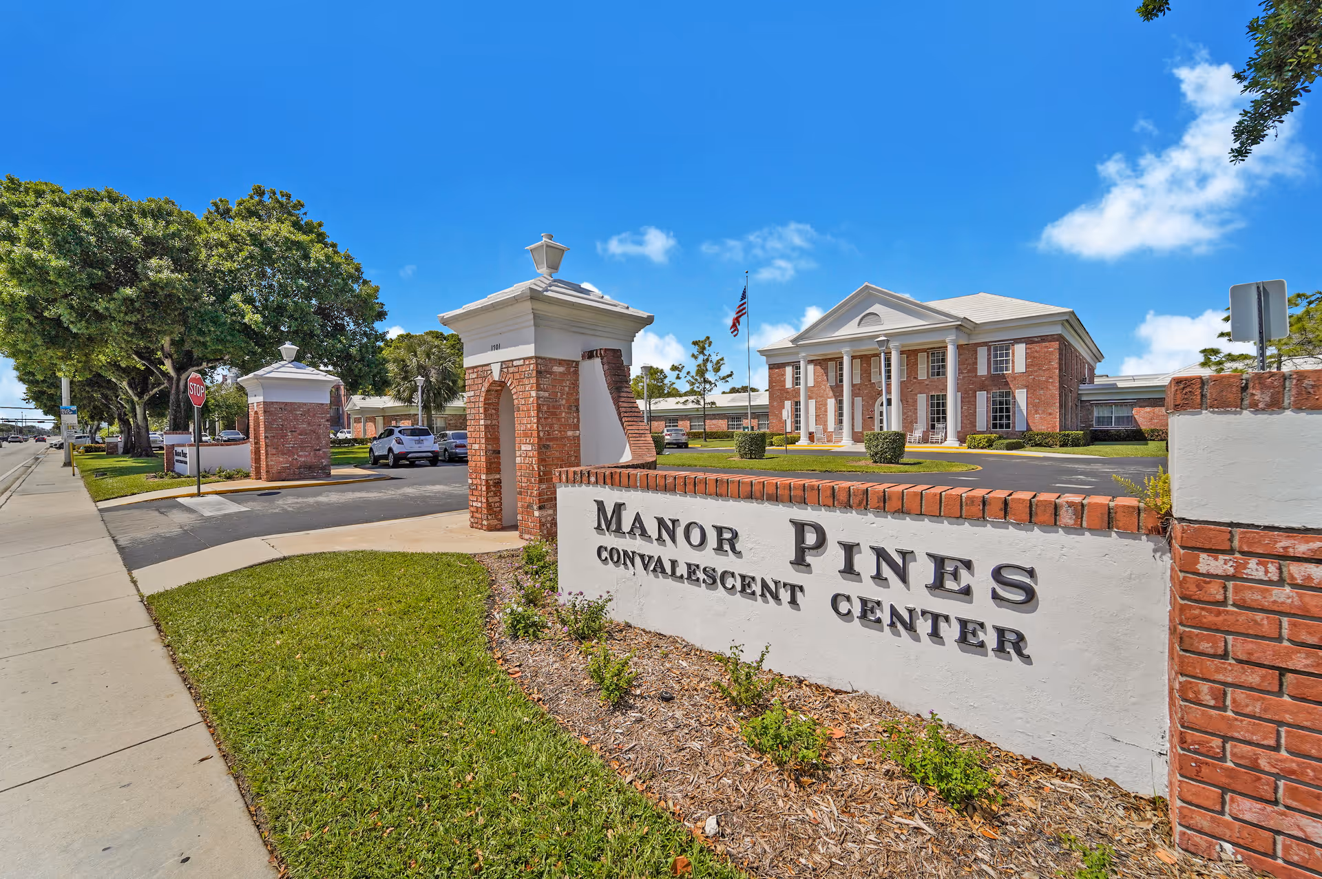 Exterior view of Manor Pines Convalescent Center showing a brick and white sign with the facility name, a driveway entrance with brick pillars, a large brick building with white columns in the background, green grass, trees, and a bright blue sky with some clouds.
